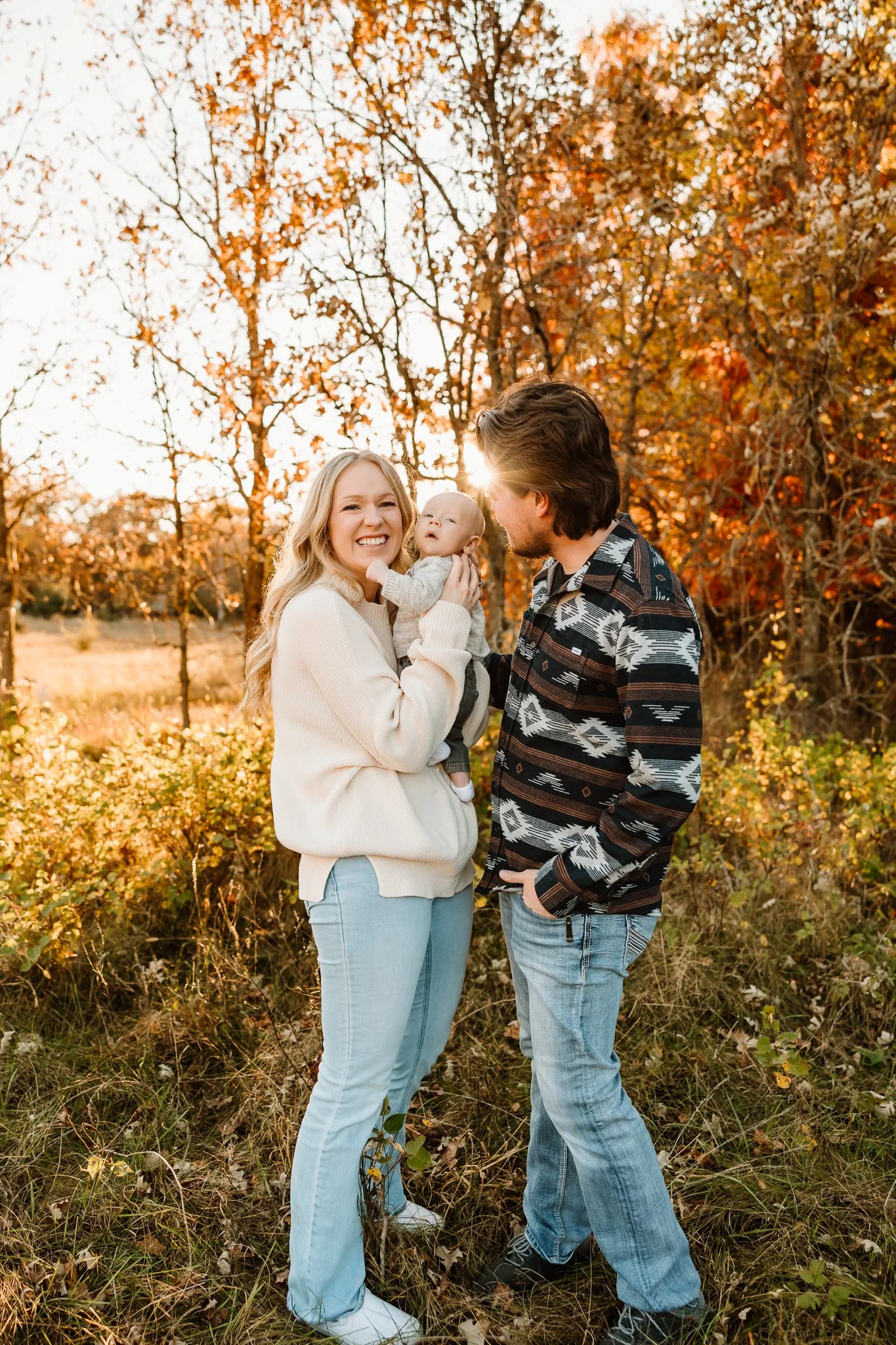 A family of three, a woman holding a baby and a man, standing outdoors in fall with orange and yellow leaves, smiling as the sun sets behind trees.