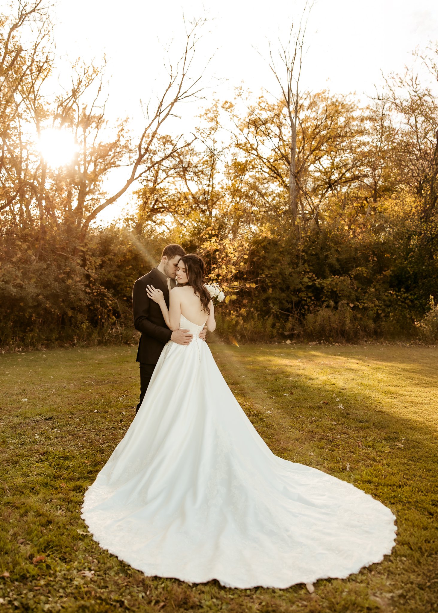 A bride and groom embracing outdoors during sunset, with the bride in a white wedding gown and the groom in a black suit, surrounded by autumn trees.
