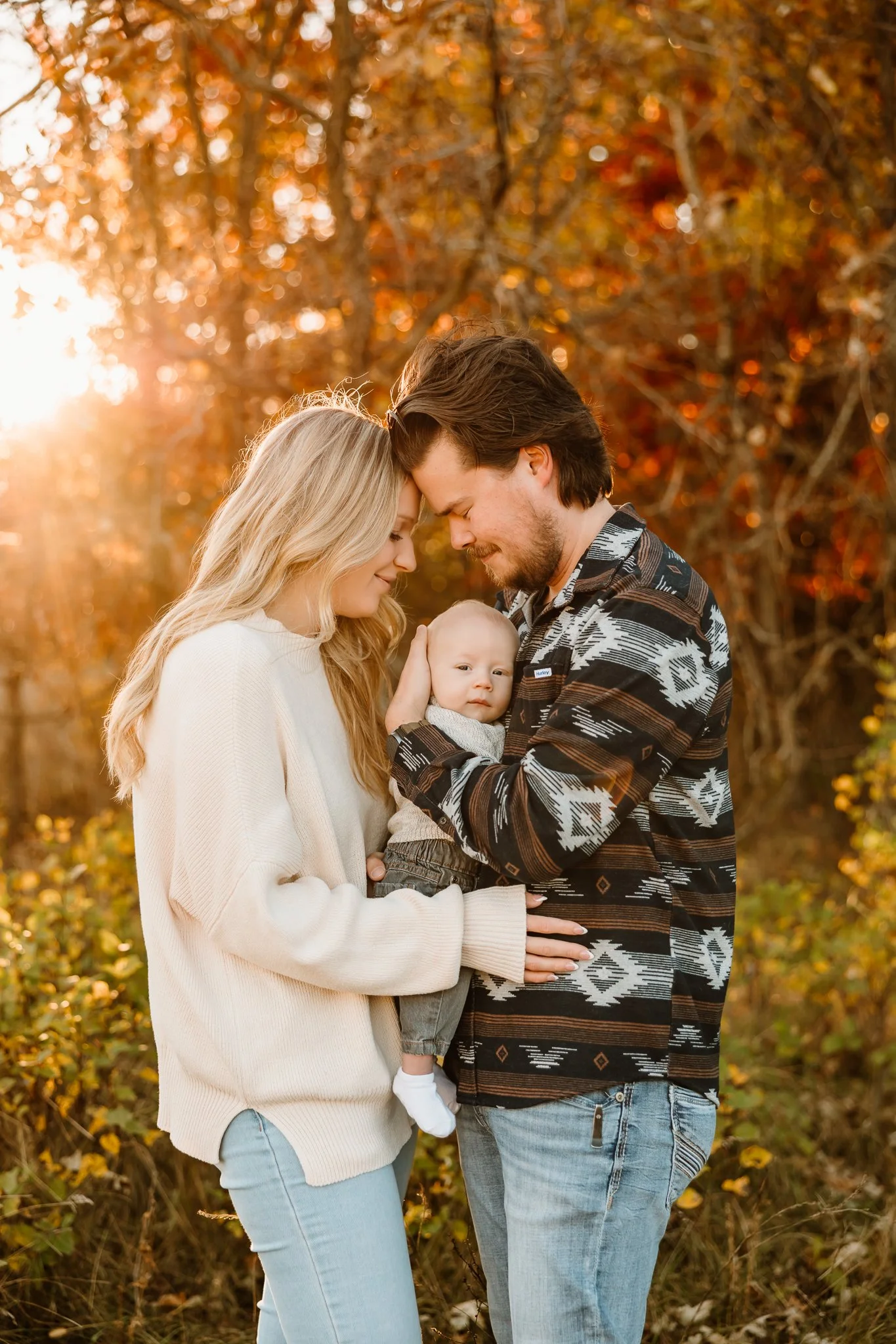 A family of three stands close together outdoors during fall, with warm sunlight filtering through orange and red leaves. The mother, with long blonde hair, gently holds their toddler, who is looking at the camera, while the father, with brown hair a