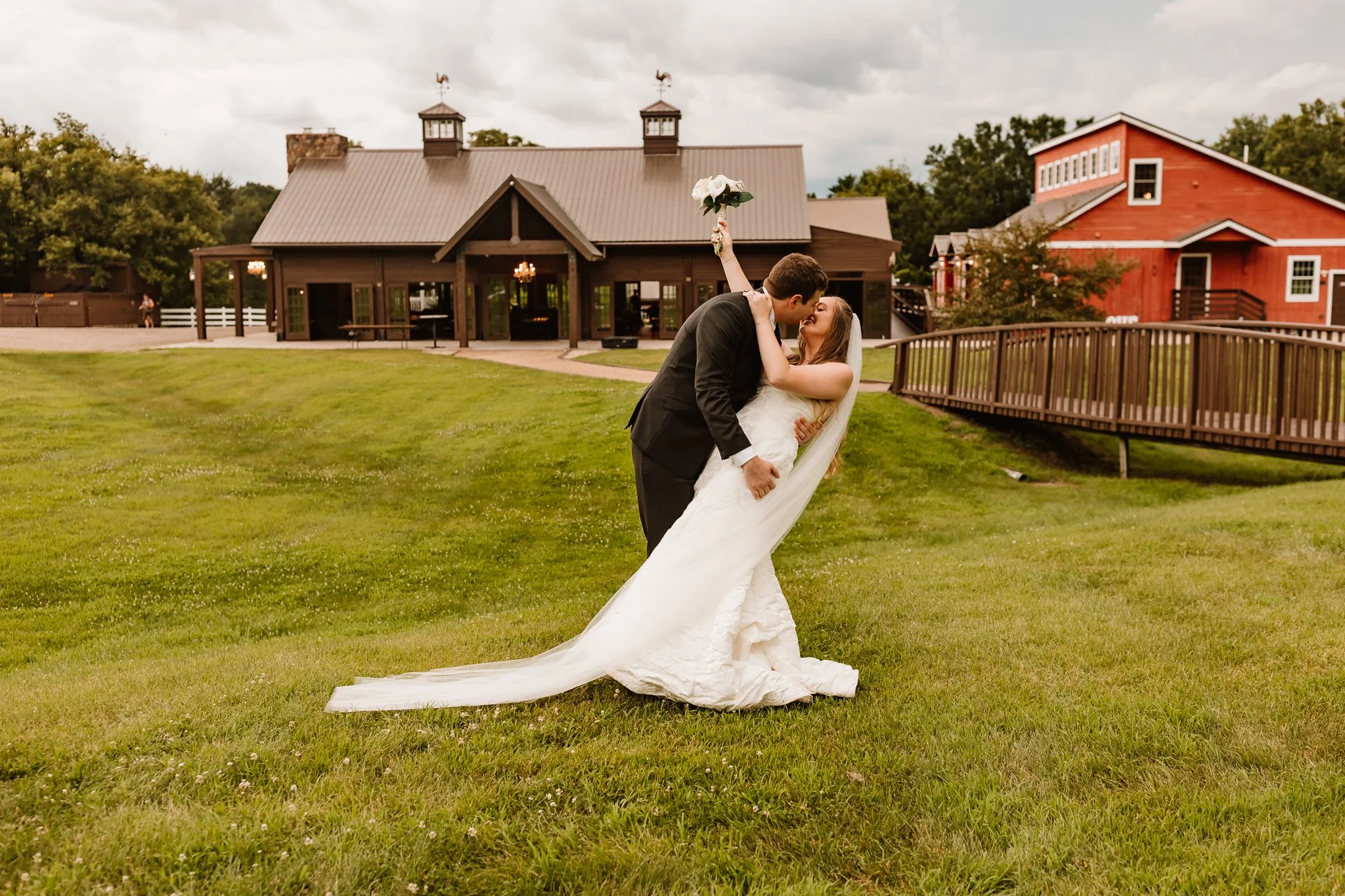 A bride and groom kissing outside on a grassy area during their wedding, with the bride holding a bouquet of white flowers and the groom in a black suit. There are buildings and a wooden fence in the background.