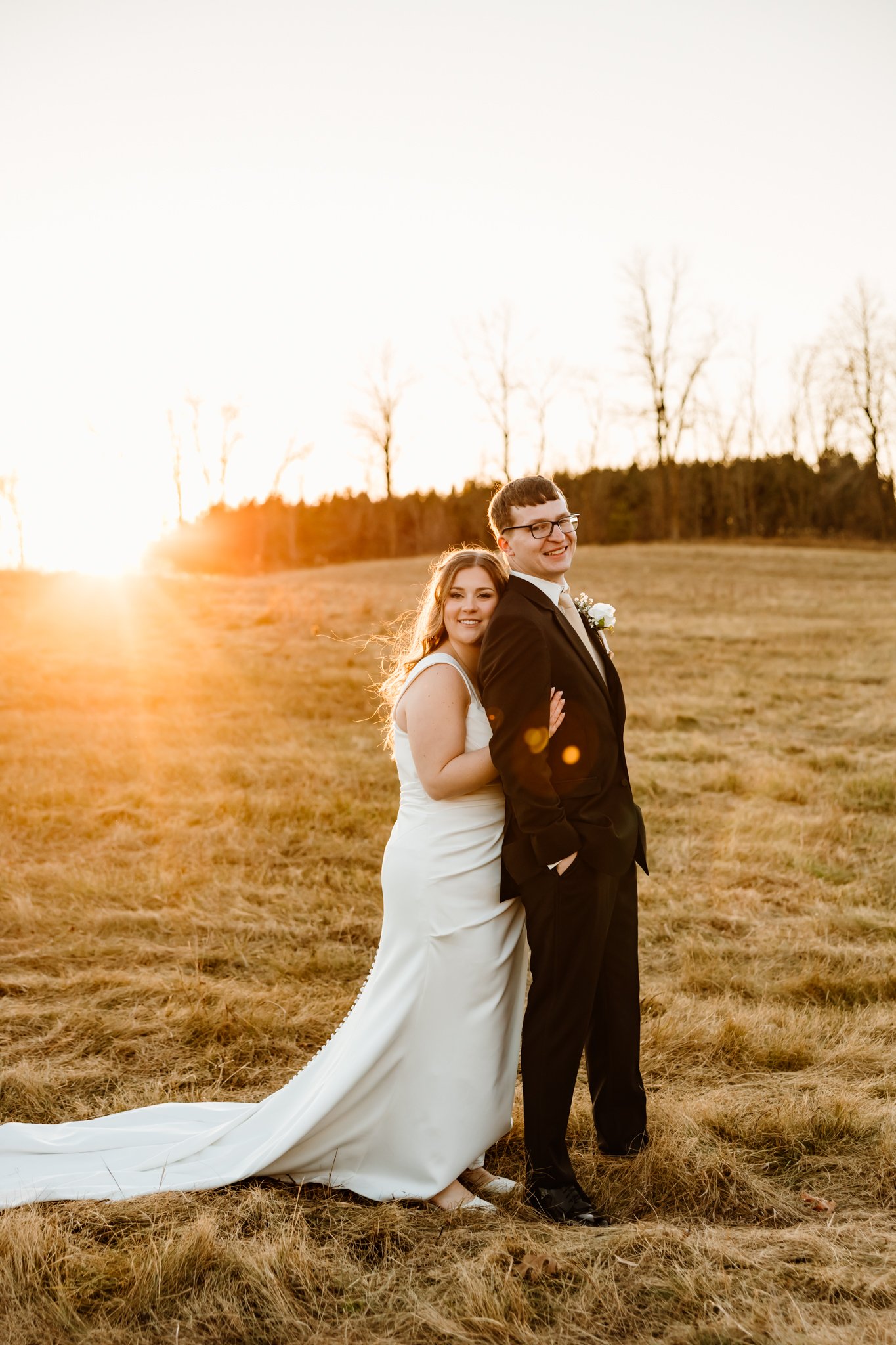 A newlywed couple standing in a field at sunset, with the bride in a white wedding gown and the groom in a black suit, smiling and holding each other.