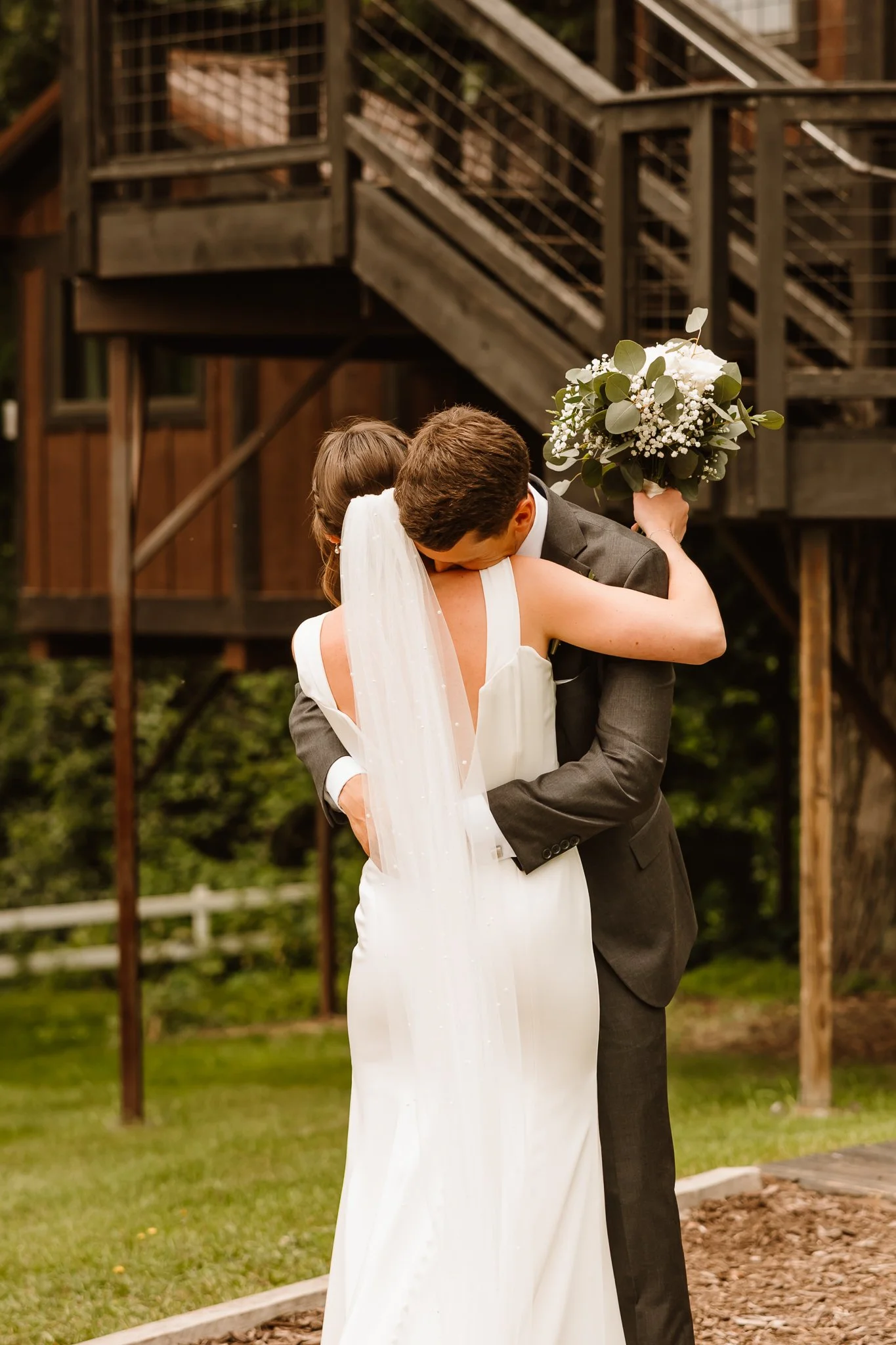 A bride and groom hugging outdoors, with the bride holding a bouquet of white flowers and greenery, near a wooden building with stairs.