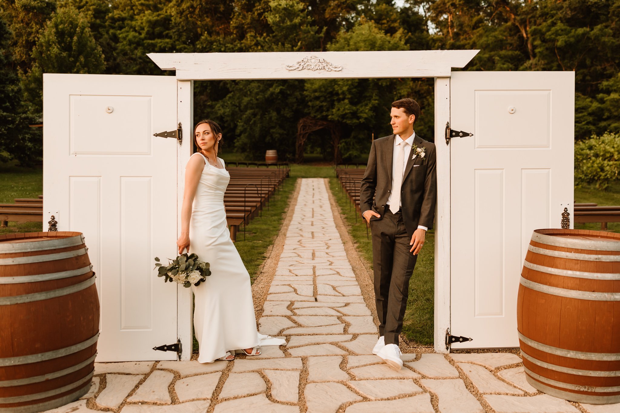 A bride and groom stand on opposite sides of a white open garden gate, with a stone pathway leading into a green outdoor area.