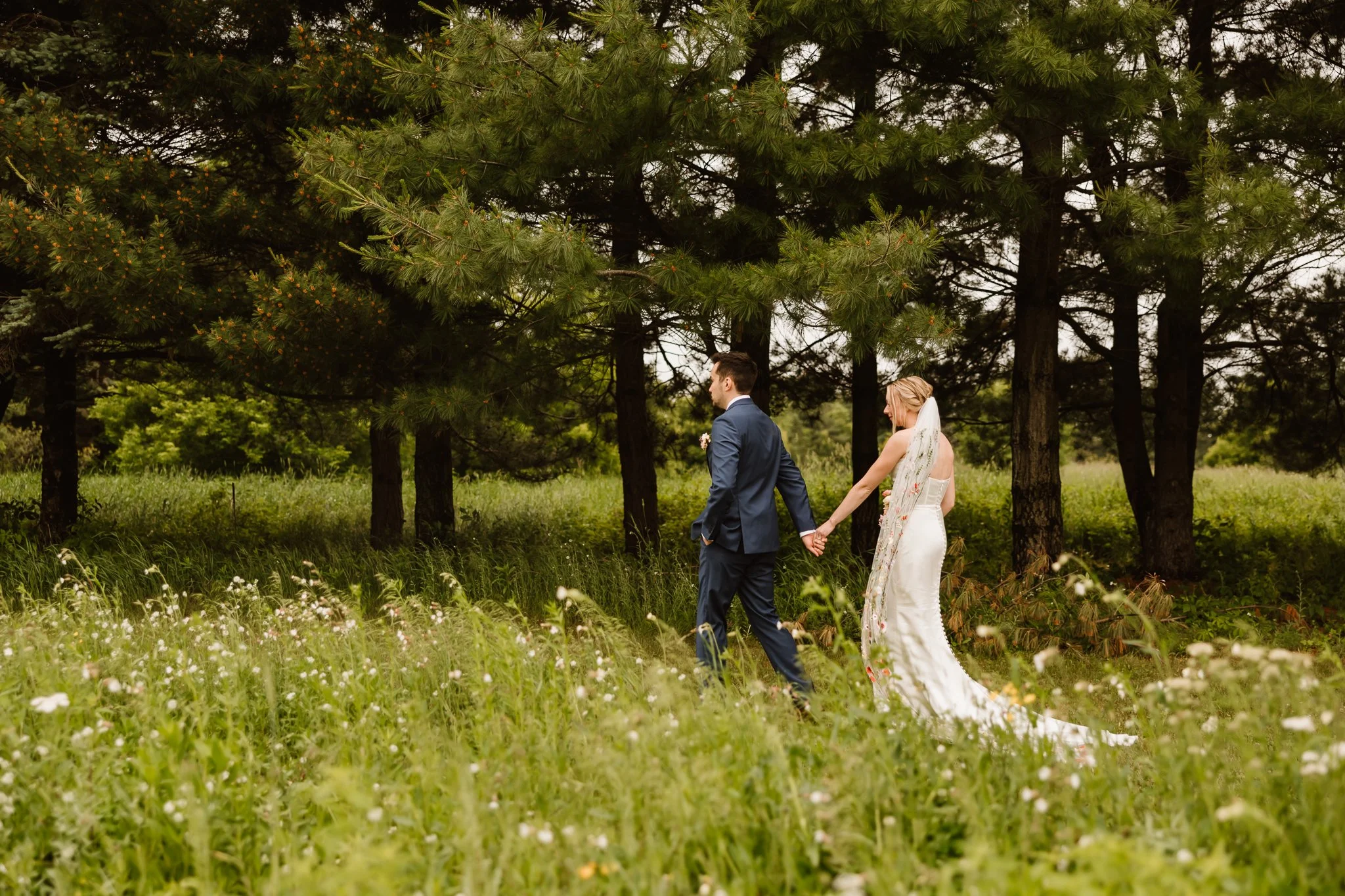 A bride and groom holding hands while walking through a grassy field with wildflowers, surrounded by tall trees in a natural outdoor setting.