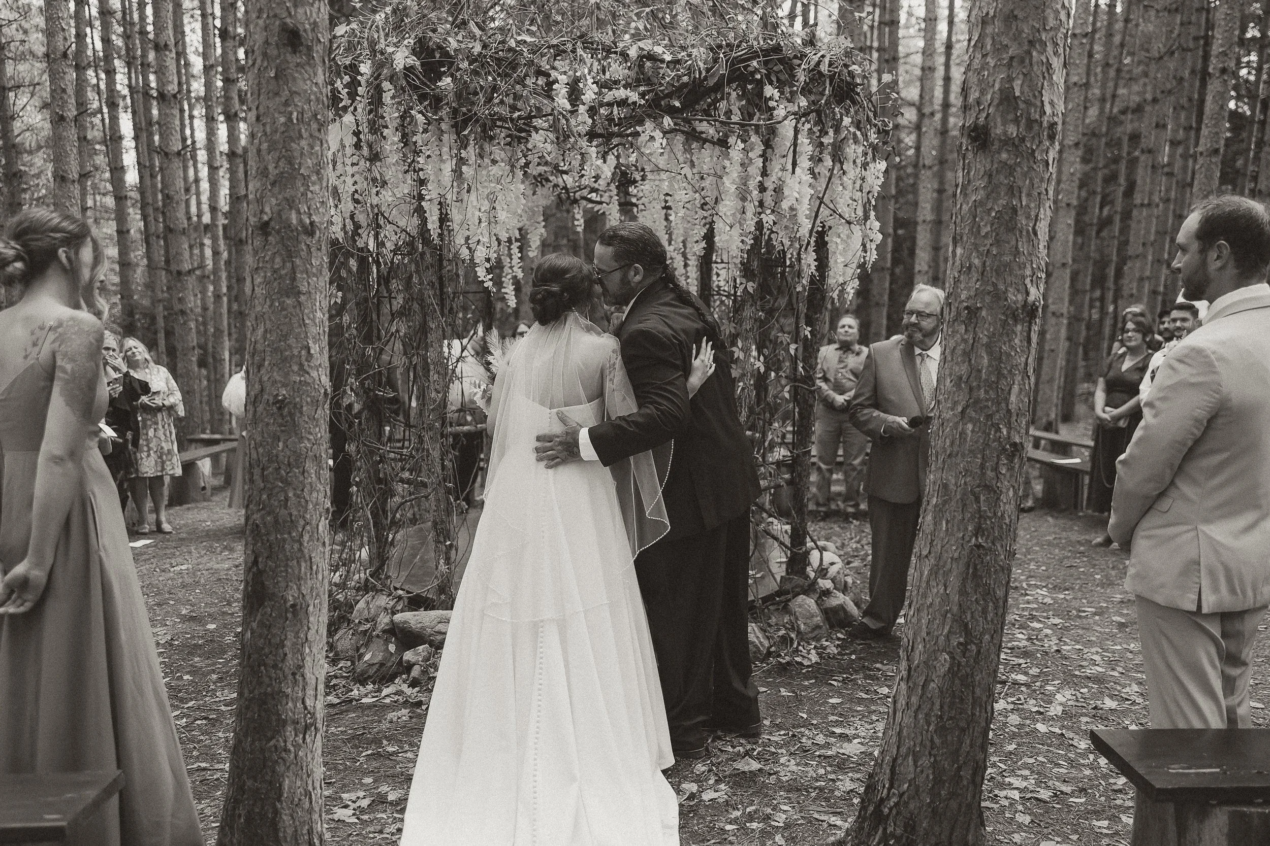 A wedding ceremony taking place in a forest, with a bride and groom sharing a kiss under an arch decorated with flowers and foliage, surrounded by guests.