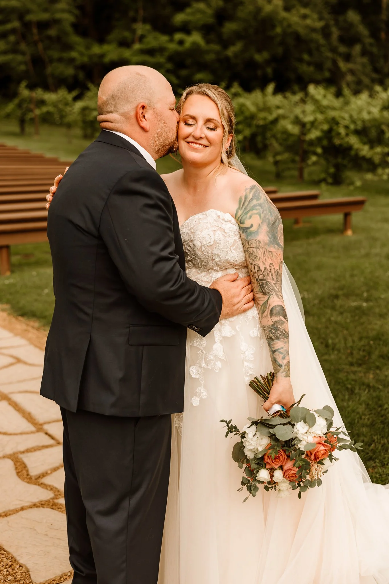 A bride and groom share a kiss outdoors on her wedding day, with the groom wearing a black suit and the bride holding a bouquet of peaches and white flowers, smiling with eyes closedHope Glen Farm Cottage Grove Minnesota 