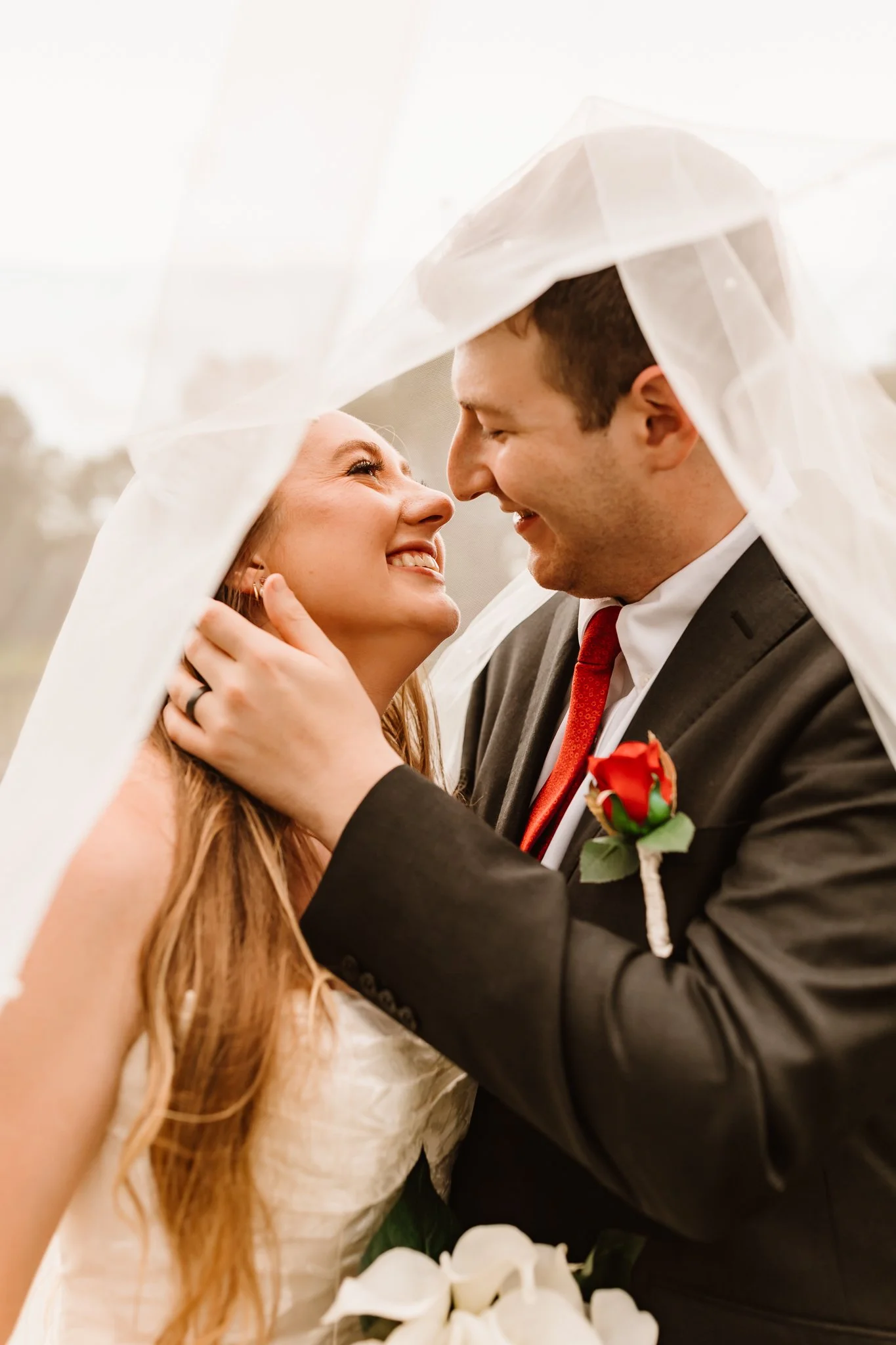 A bride and groom on their wedding day, smiling and gazing into each other's eyes, under a sheer white veil.