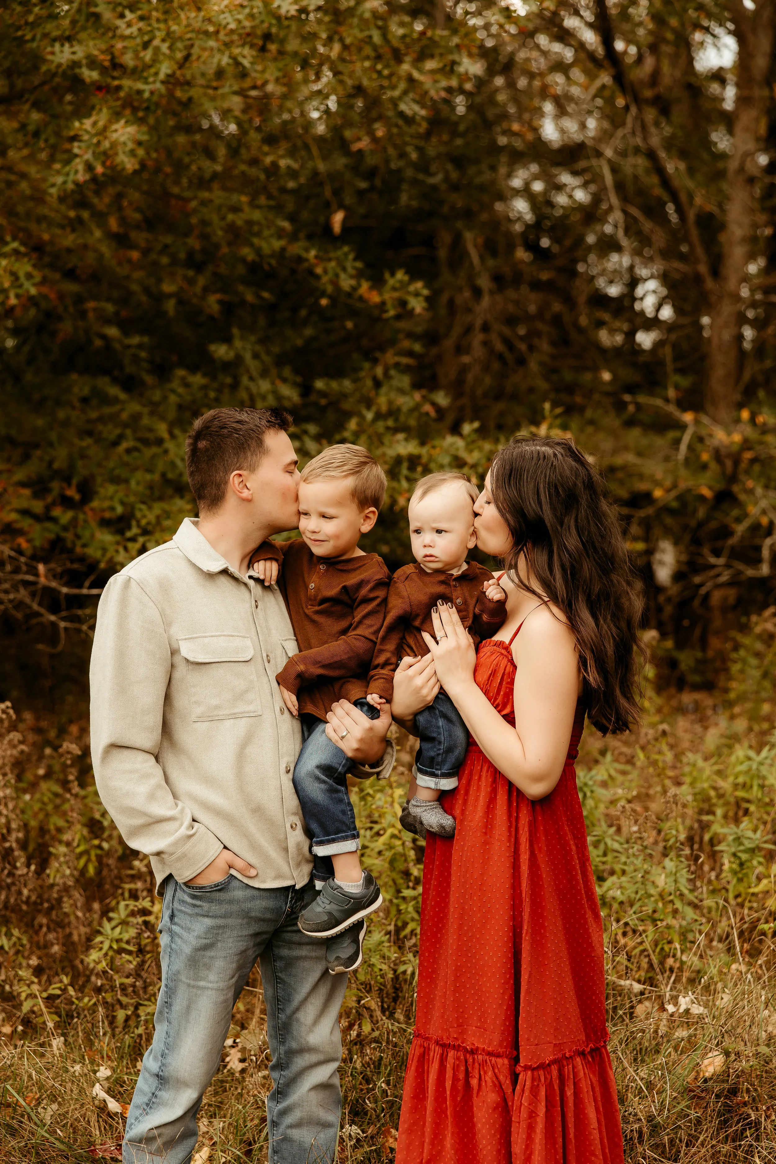 Family of four outdoors during autumn; father and mother with two young boys, the father kissing one boy and the mother kissing the other in a forested area. Minnesota family photographer 