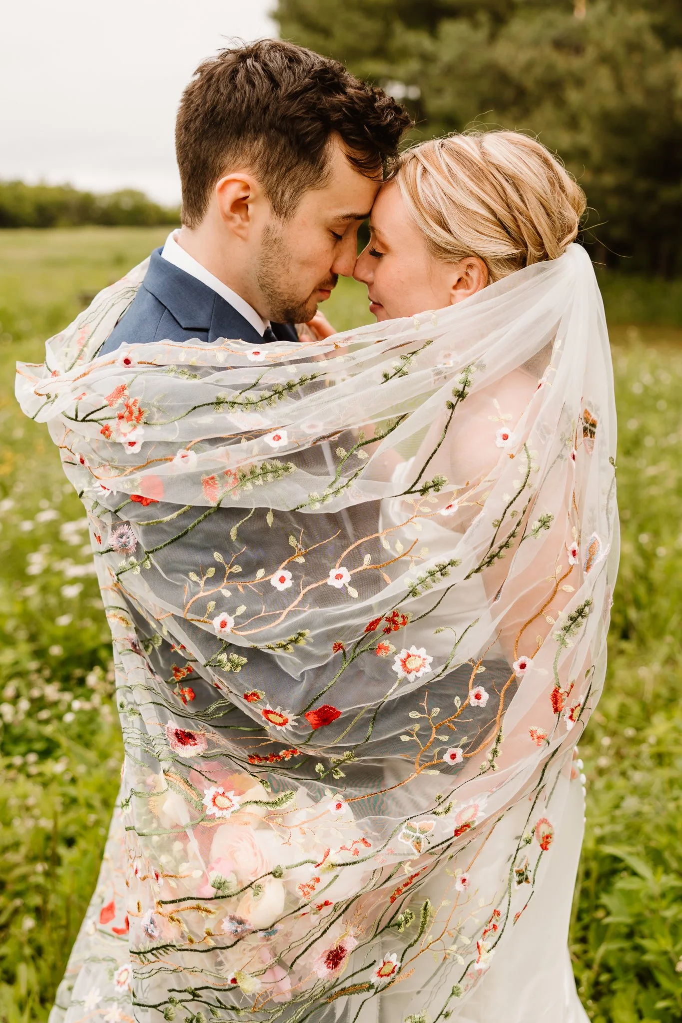 A bride and groom with foreheads touching under a sheer, embroidered floral veil in a green outdoor field.