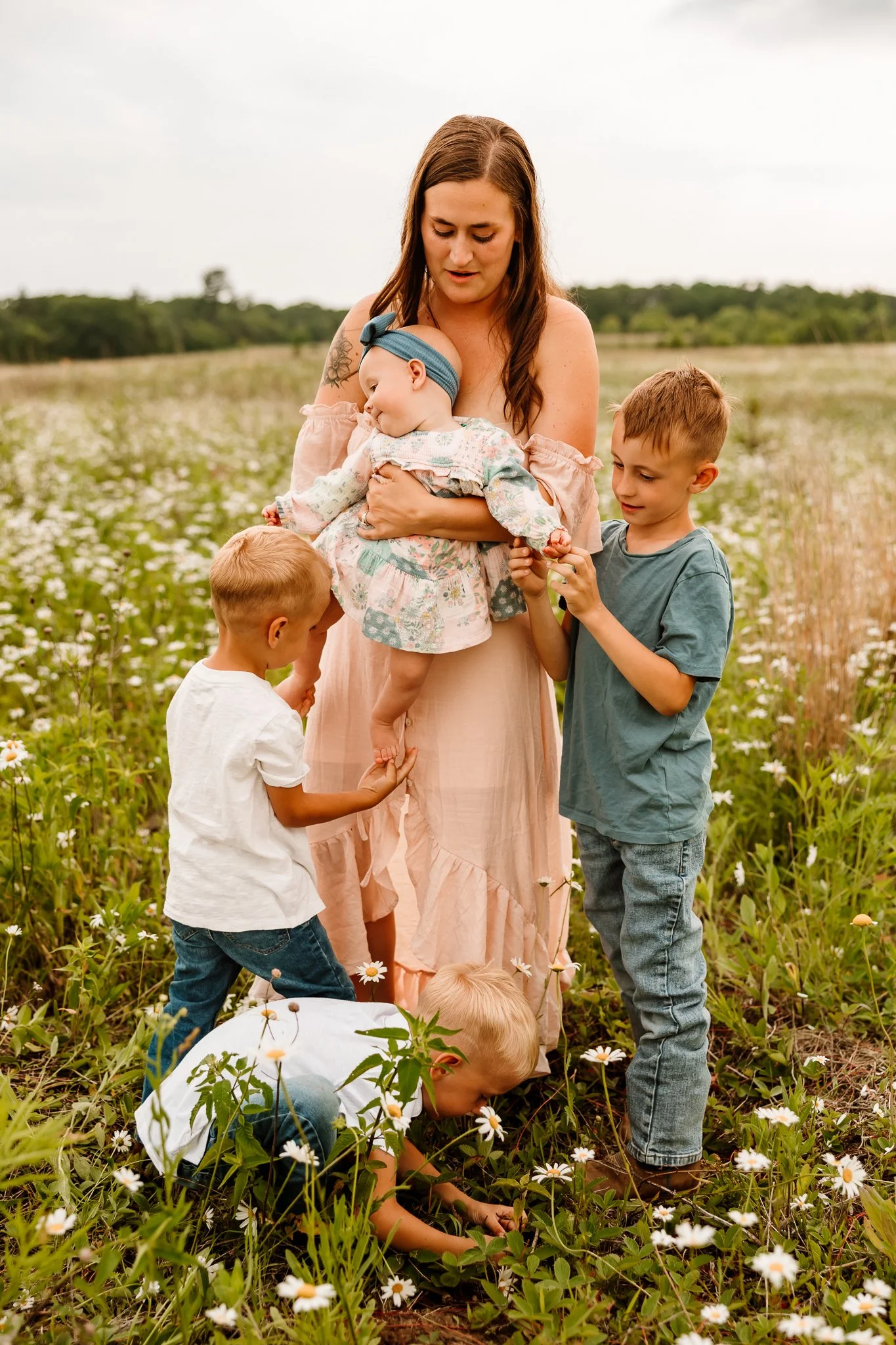 A woman with long brown hair wearing a pink dress stands in a field of daisies, holding a young girl in a floral dress and blue headband, while four young boys, dressed casually, surround her and look at the girl or the flowers, with a green landscap