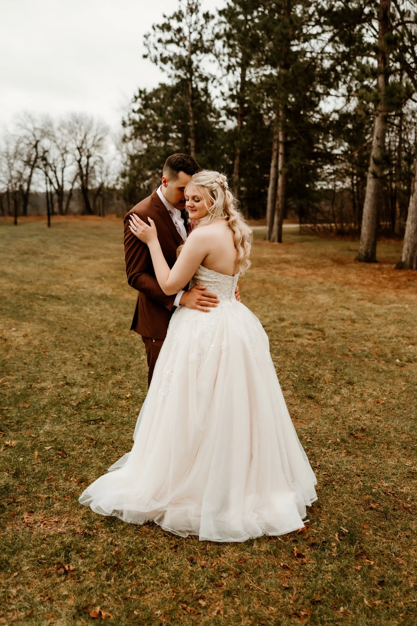 A bride and groom embrace outdoors in a grassy area with trees, dressed in wedding attire, during daytime.