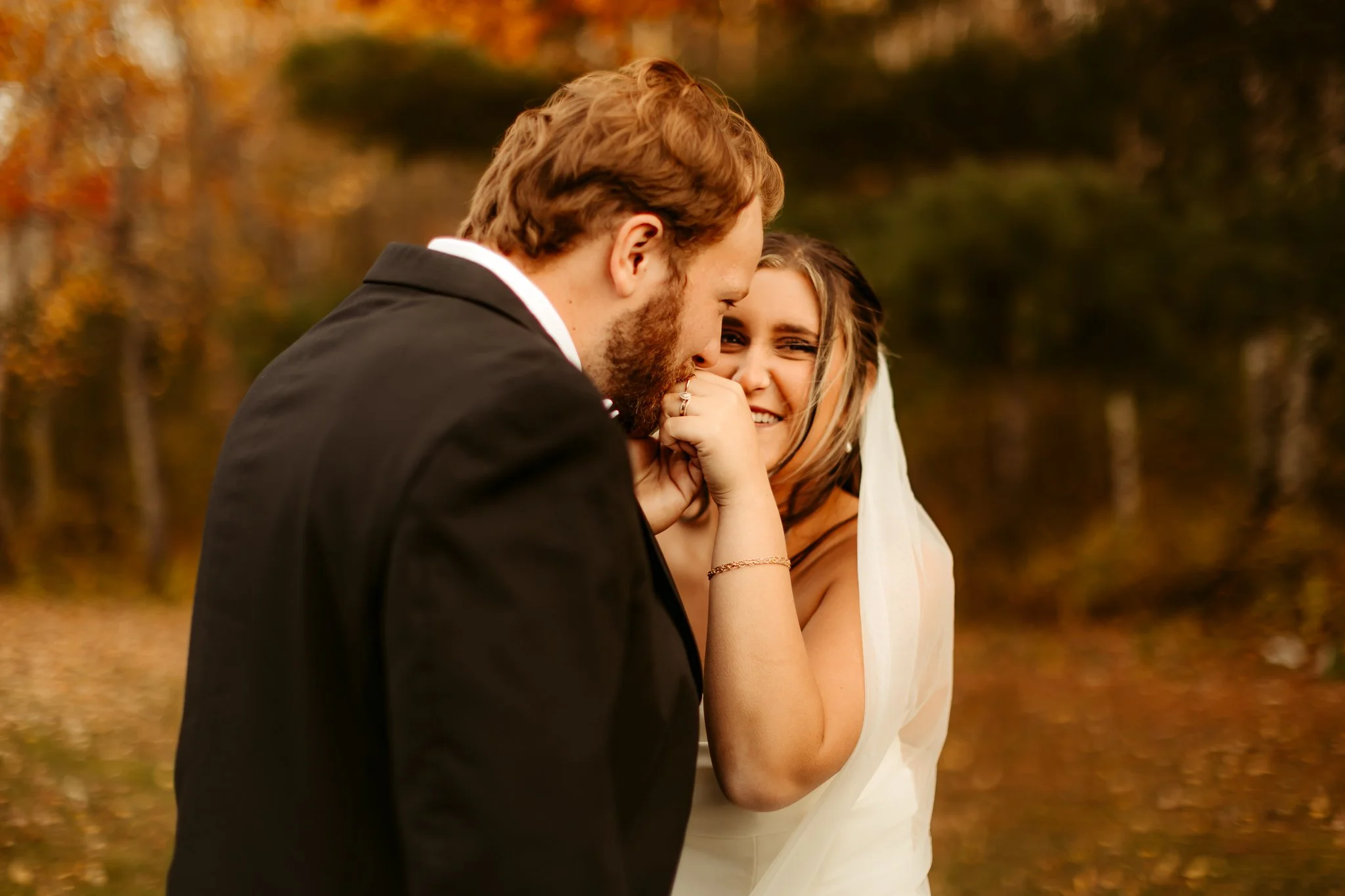 A bride and groom share a tender moment outdoors during autumn, with the bride smiling and holding the groom's hand close to her face.