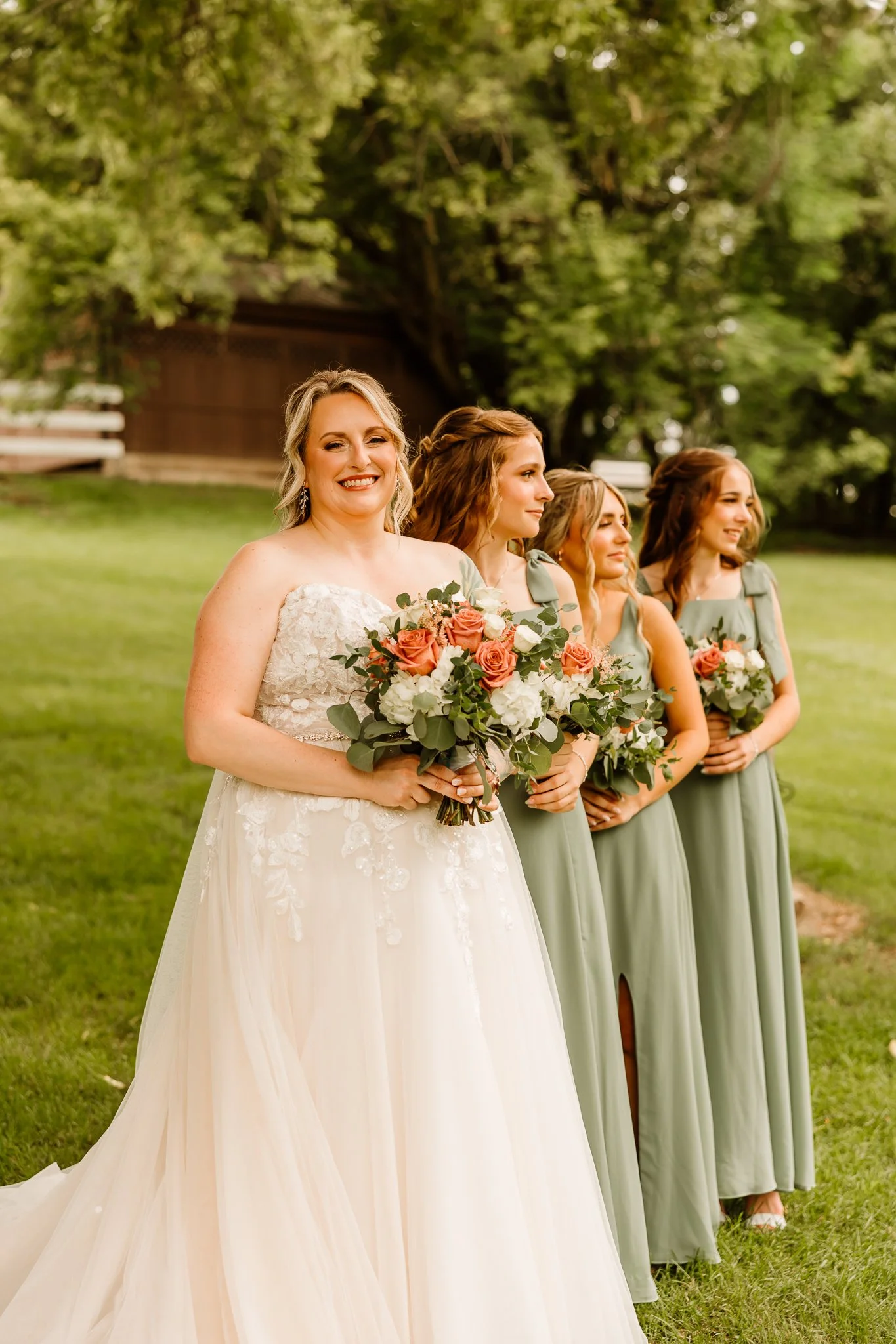 A bride in a white wedding gown holding a bouquet of pink and white roses, standing outdoors with three bridesmaids in sage green dresses holding similar bouquets, green trees in the background.