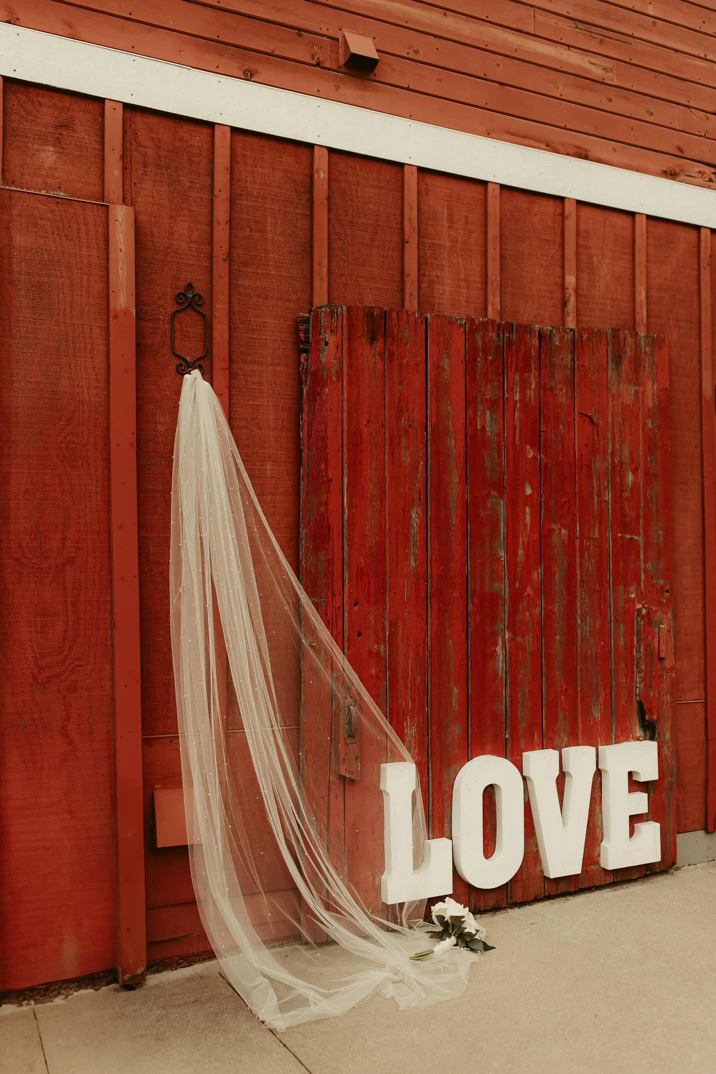 Decorative red wooden wall with large white letters spelling 'LOVE', a white veil hanging on a black hook, and a small bouquet of white roses on the ground.