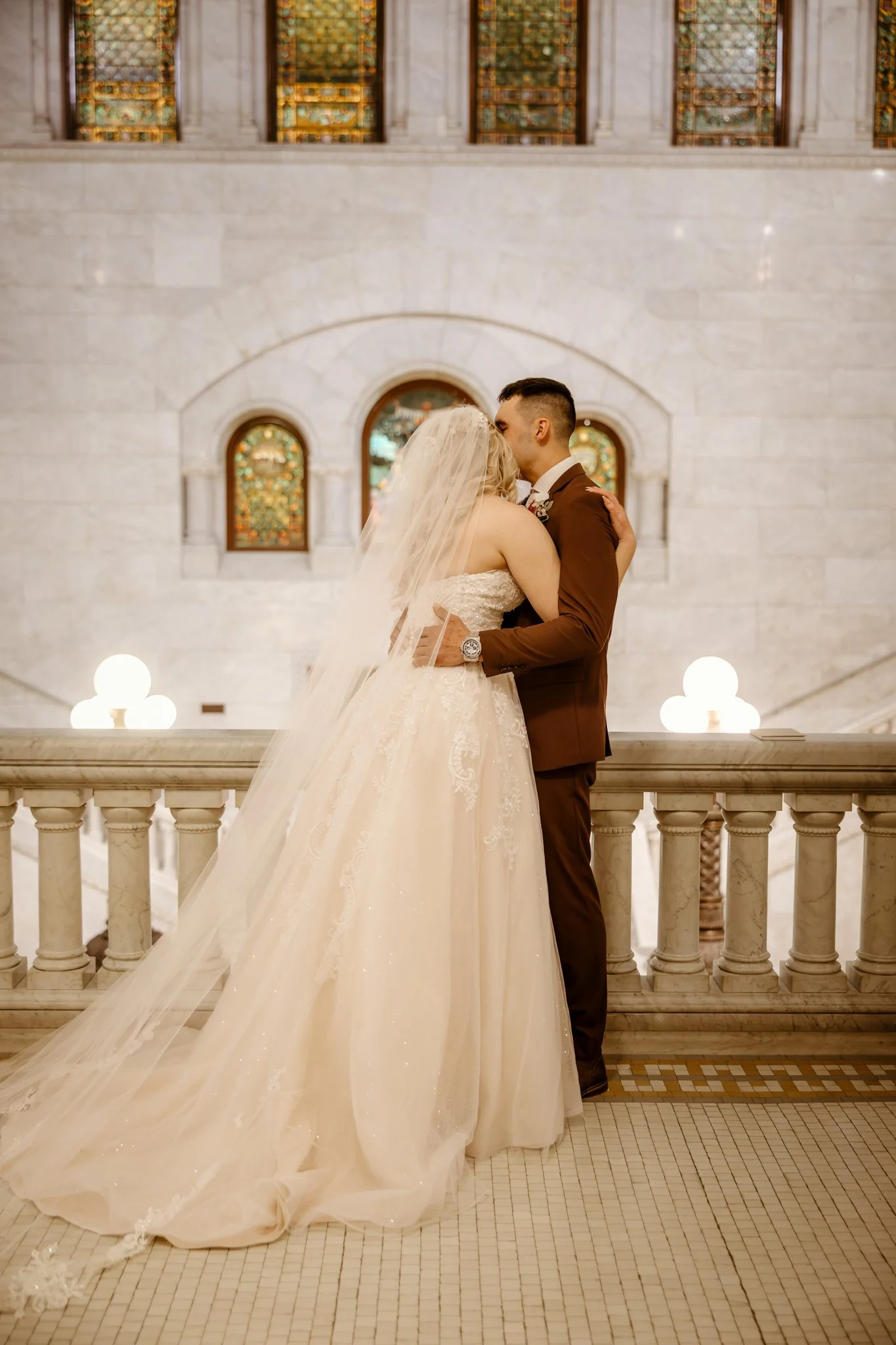 A bride and groom sharing a kiss in a church or grand hall, with stained glass windows and marble architecture in the background.