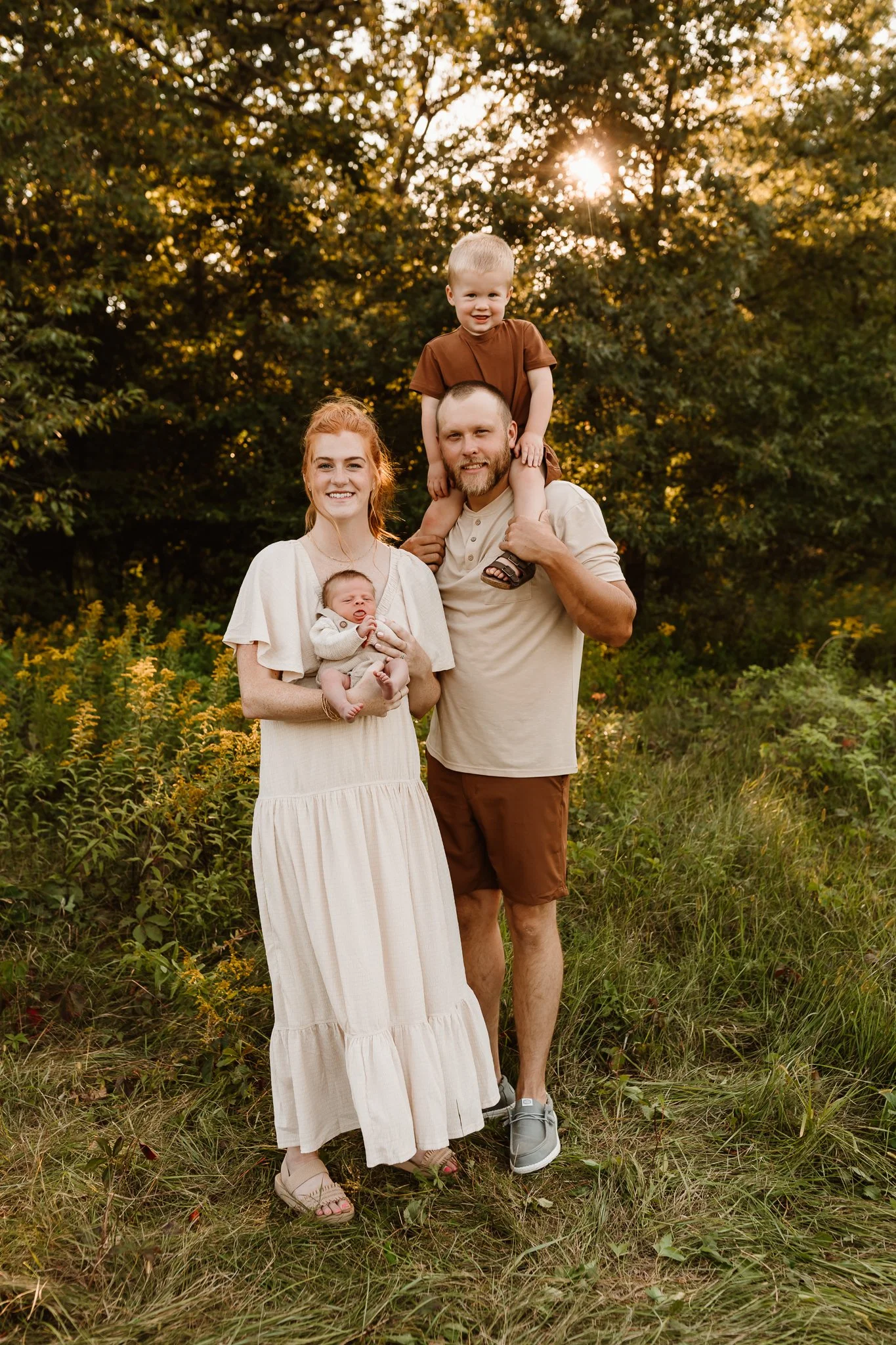 A family of four outdoors during sunset, with trees and greenery in the background. The mother is holding a baby, the father is carrying a young boy on his shoulders, all smiling.