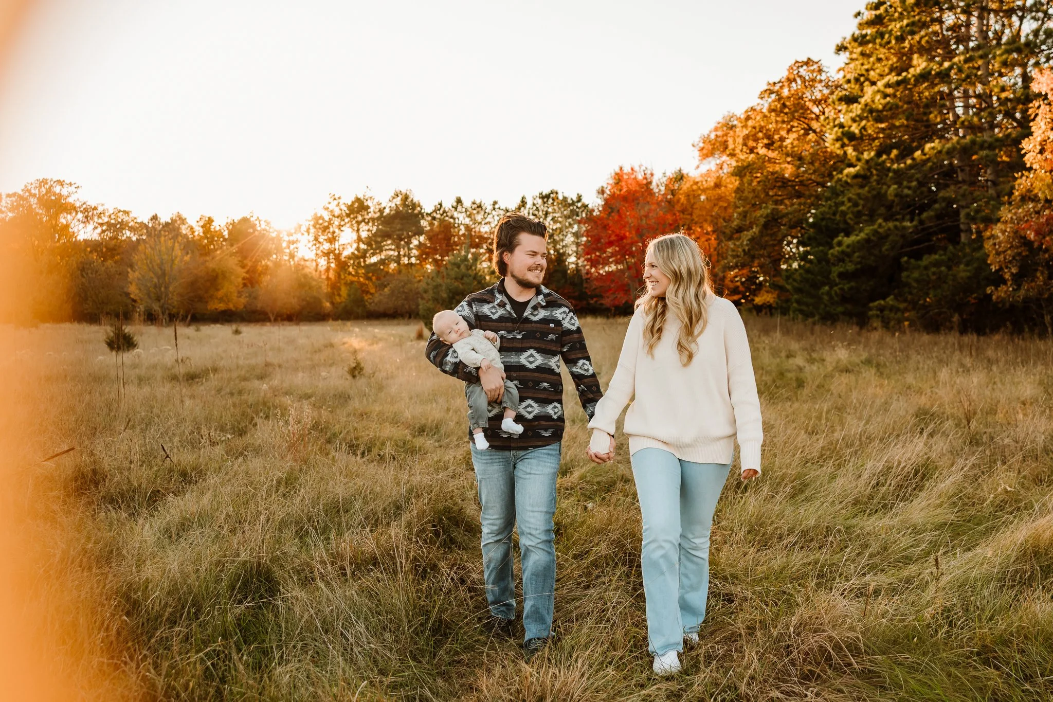 A family of three walking through a grassy field during sunset, with autumn trees in the background. The father is holding a baby, and they are holding hands.