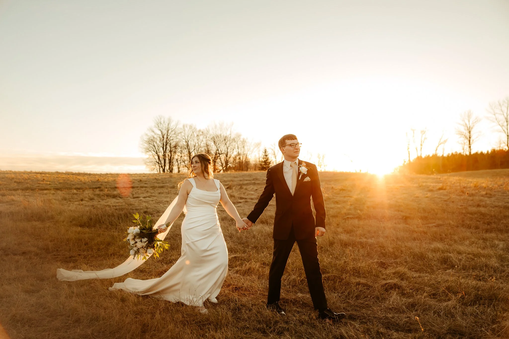 A bride and groom walking hand in hand in an open field at sunset, with the bride in a white gown holding a bouquet and the groom in a dark suit.