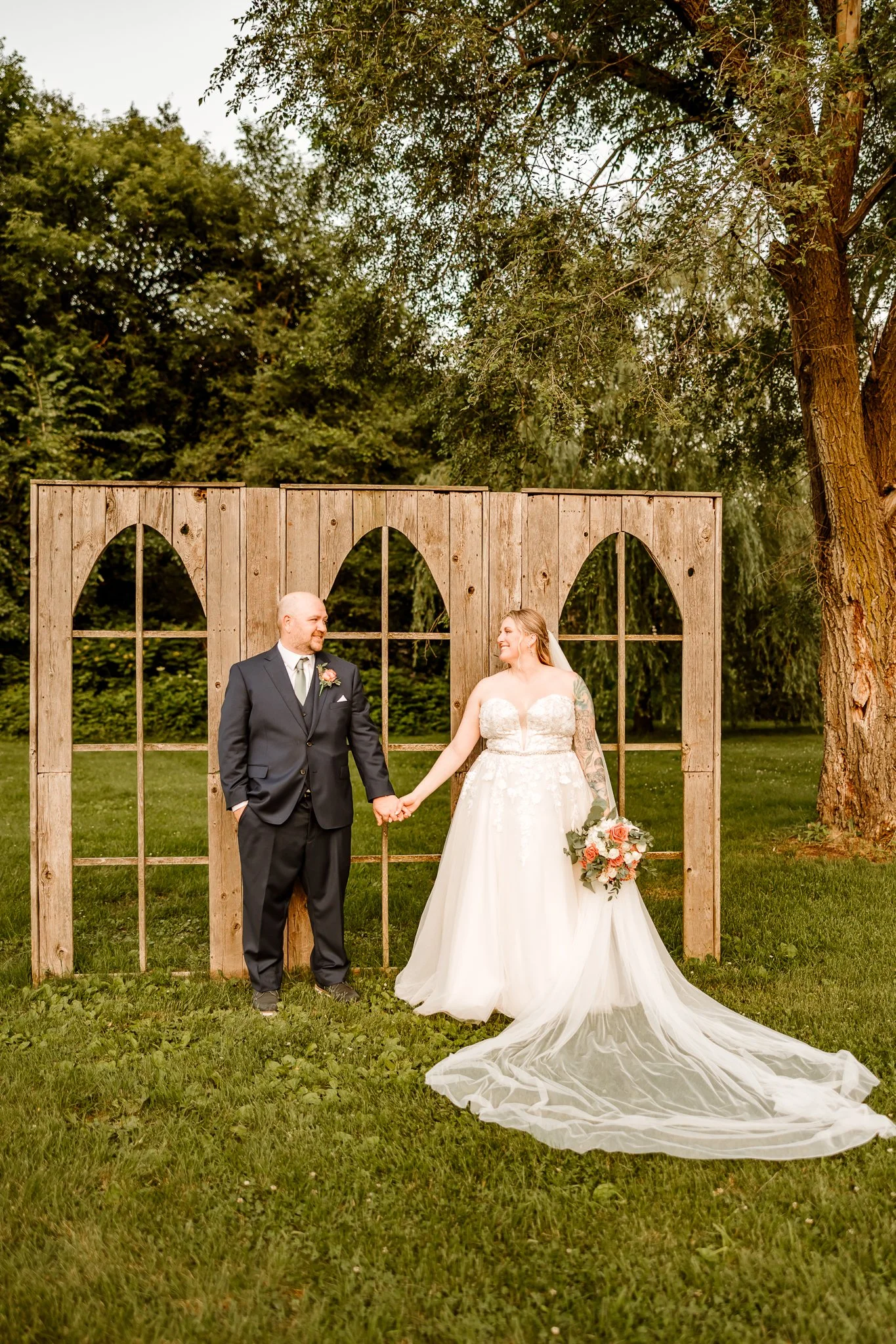 A bride and groom holding hands and smiling at each other outdoors during daylight, with a rustic wooden backdrop and green trees in the background.