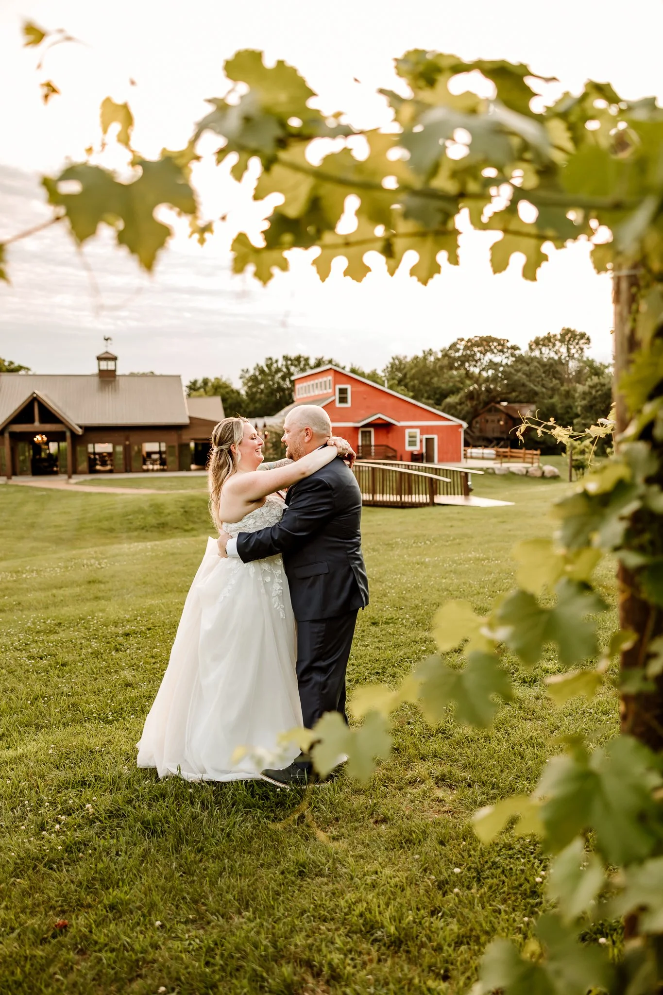 A newlywed couple in wedding attire sharing a dance on a green lawn, framed by leafy branches in a rustic outdoor setting with a barn and other farm buildings in the background during sunsetHope Glen Farm Cottage Grove Minnesota 
