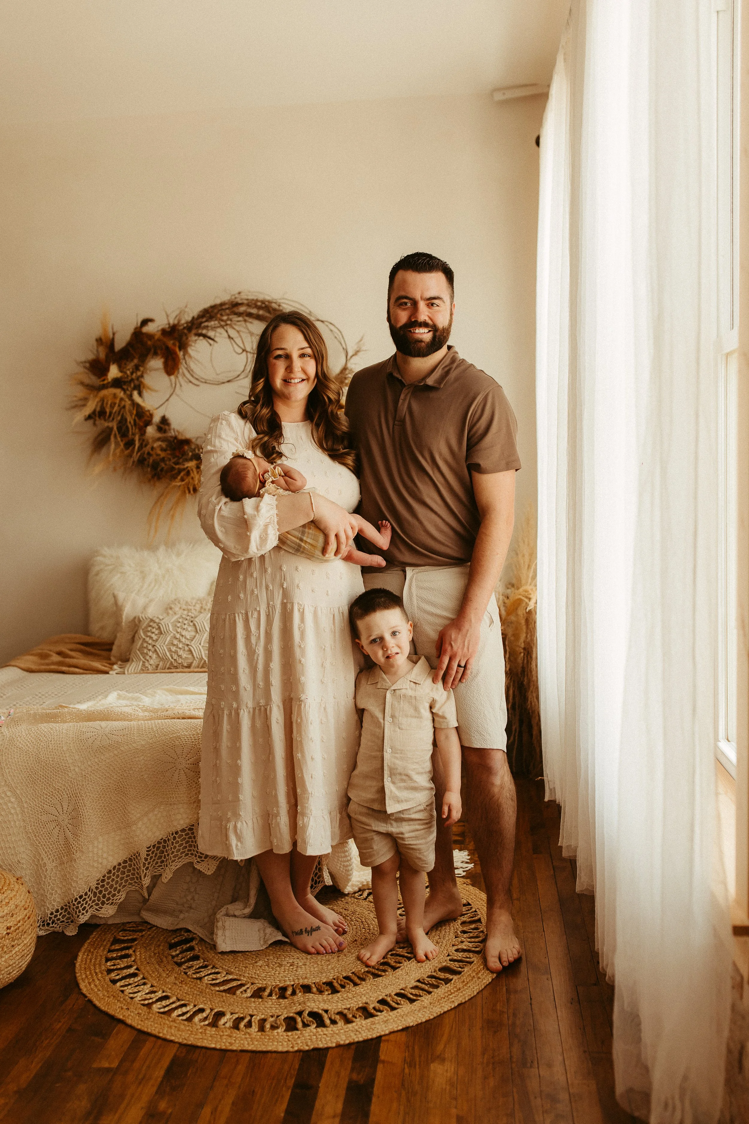 A family of four, including a mother holding a baby, father, and young son, standing in a cozy, warmly decorated bedroom with natural light, a bed with neutral-colored bedding, and bohemian-style decor in the background.
