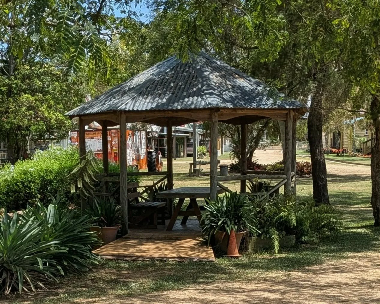 Wooden gazebo with metal roof in a park surrounded by trees and potted plants.