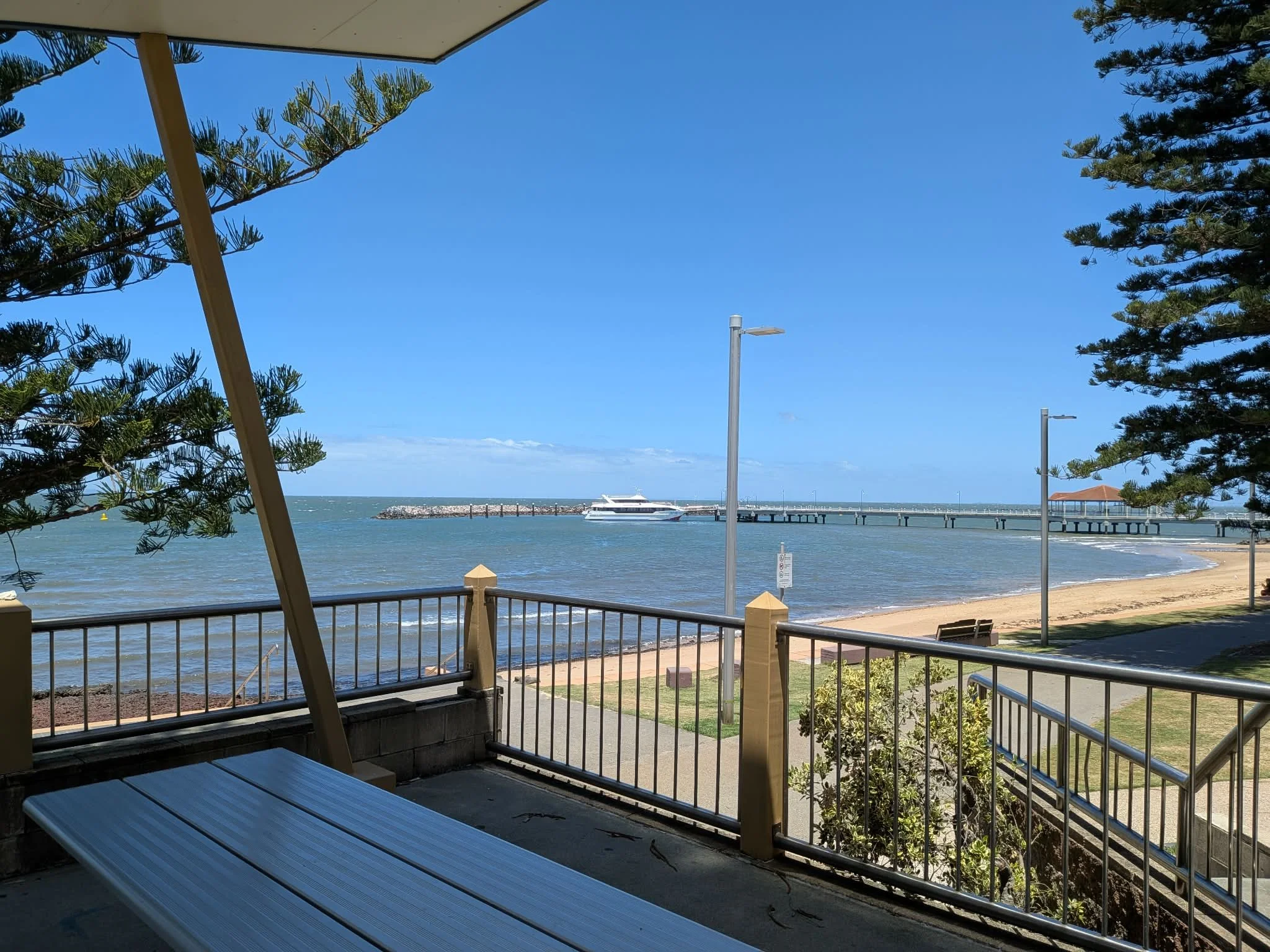 View of a beach with a pier, boats, and clear sky, seen from a shaded balcony with trees, railing, and benches. Saying goodbye on the beach.