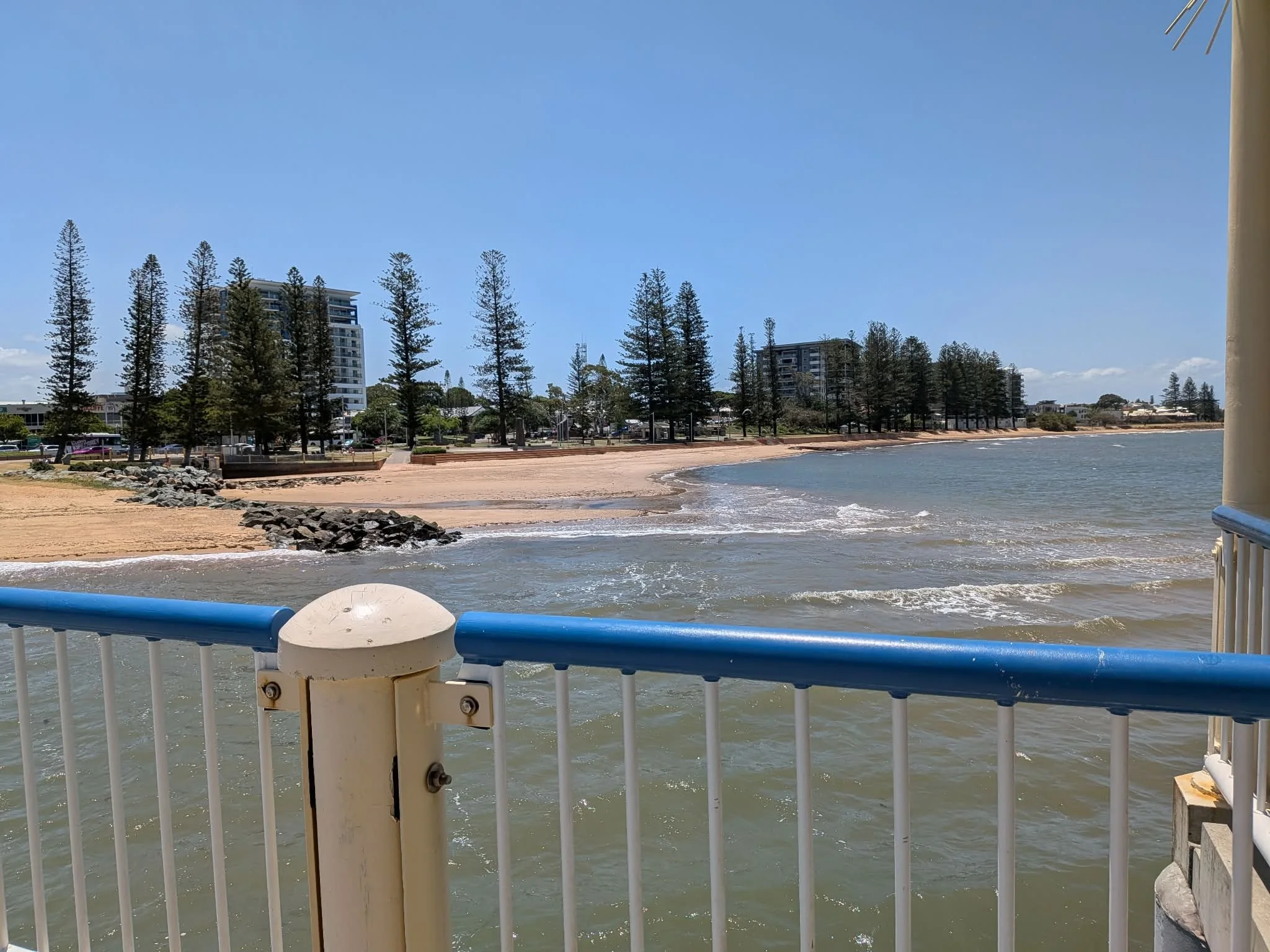 View of a beach from a pier with a metal railing, sandy shoreline, small waves, sparse trees, and buildings in the background under a blue sky. Redcliffe, Redcliffe Park, Woody Point.