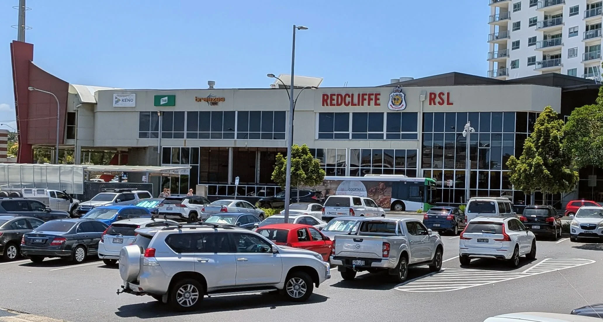 Parking lot filled with various cars in front of a building labeled 'Redcliffe RSL' with several business signs, three trees, and a taller apartment building in the background.