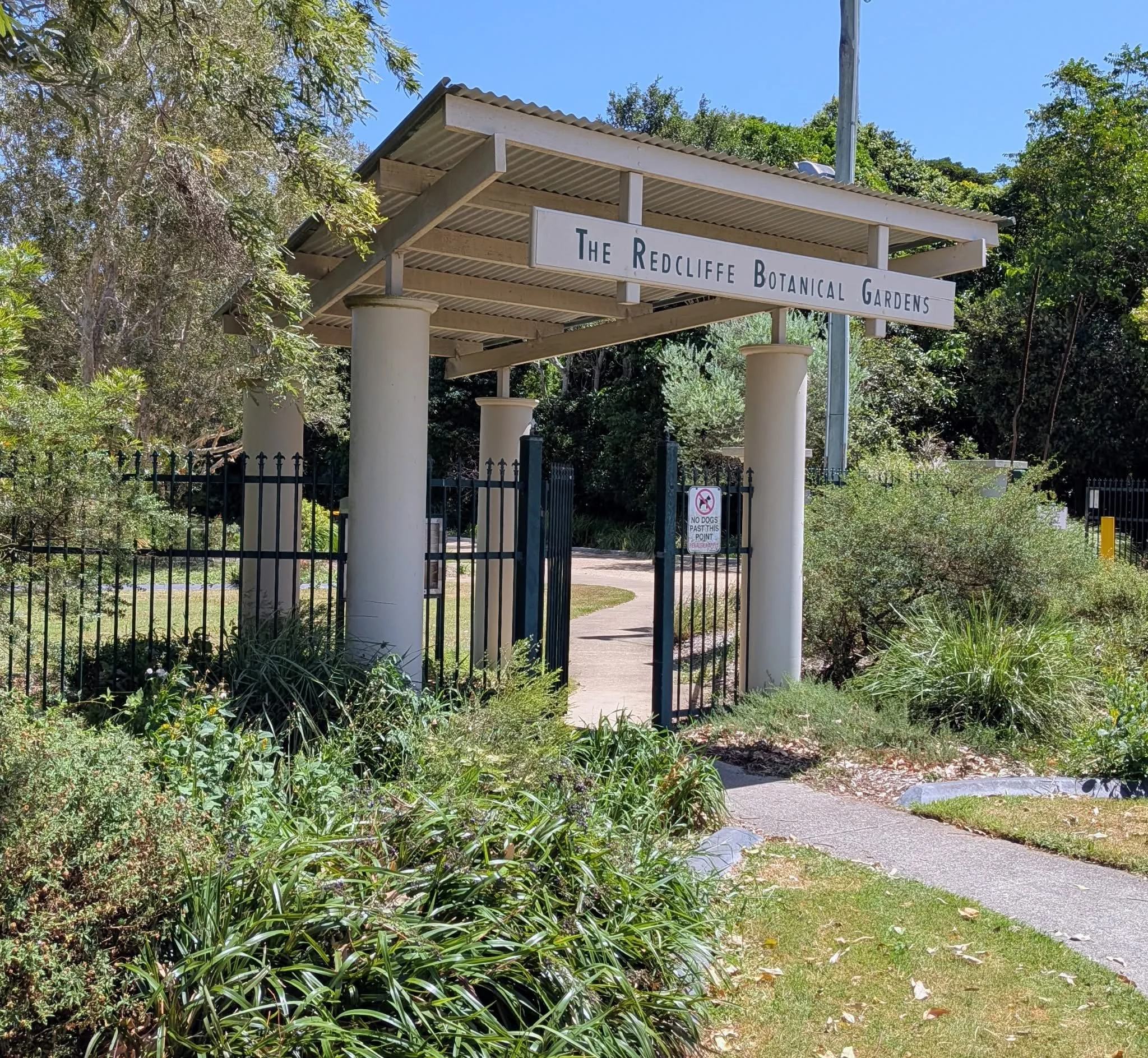 Entrance to The Redcliffe Botanical Gardens with a sign overhead, surrounded by greenery and a pathway leading inside. Redcliffe Botanical Gardens.