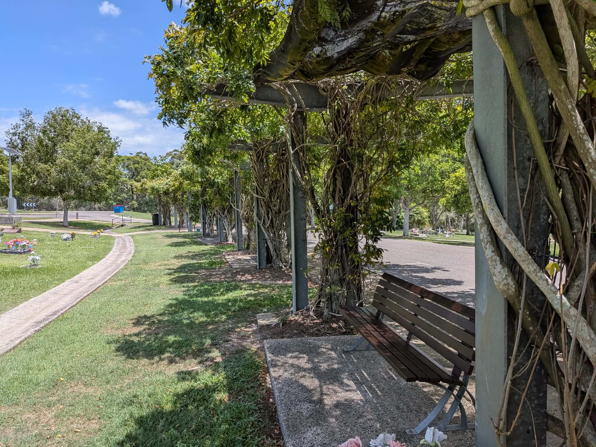 A shaded park bench under a wooden structure entwined with vines, next to a curved walking path, grassy area with flower arrangements, trees, and a blue sky with clouds. Great Northern Garden of Remembrance.