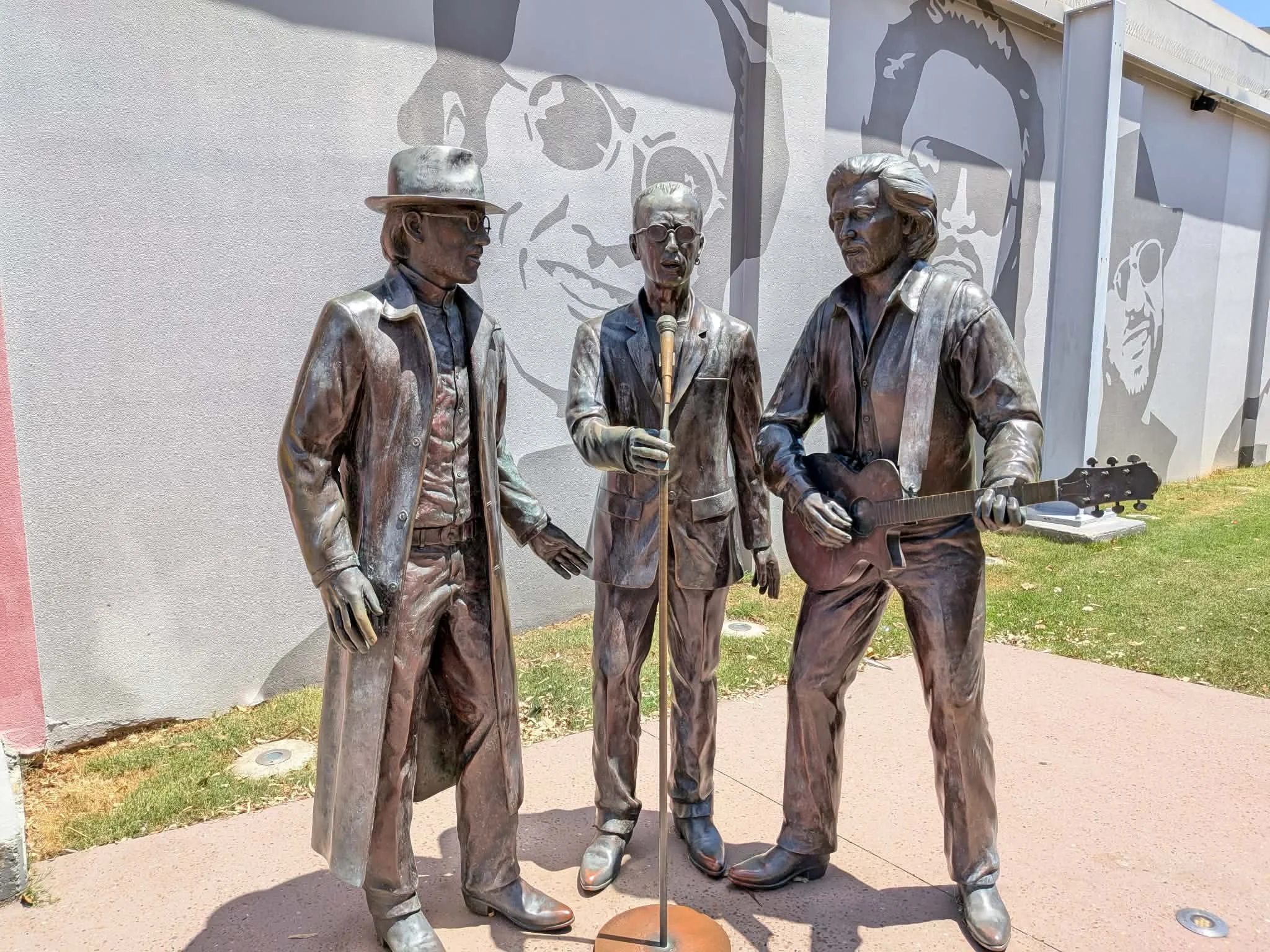 Bronze statues of three men, one with a hat and glasses, another holding a microphone, and the third playing an acoustic guitar, against a wall with a large face mural. Recliffe, Bee Gees.