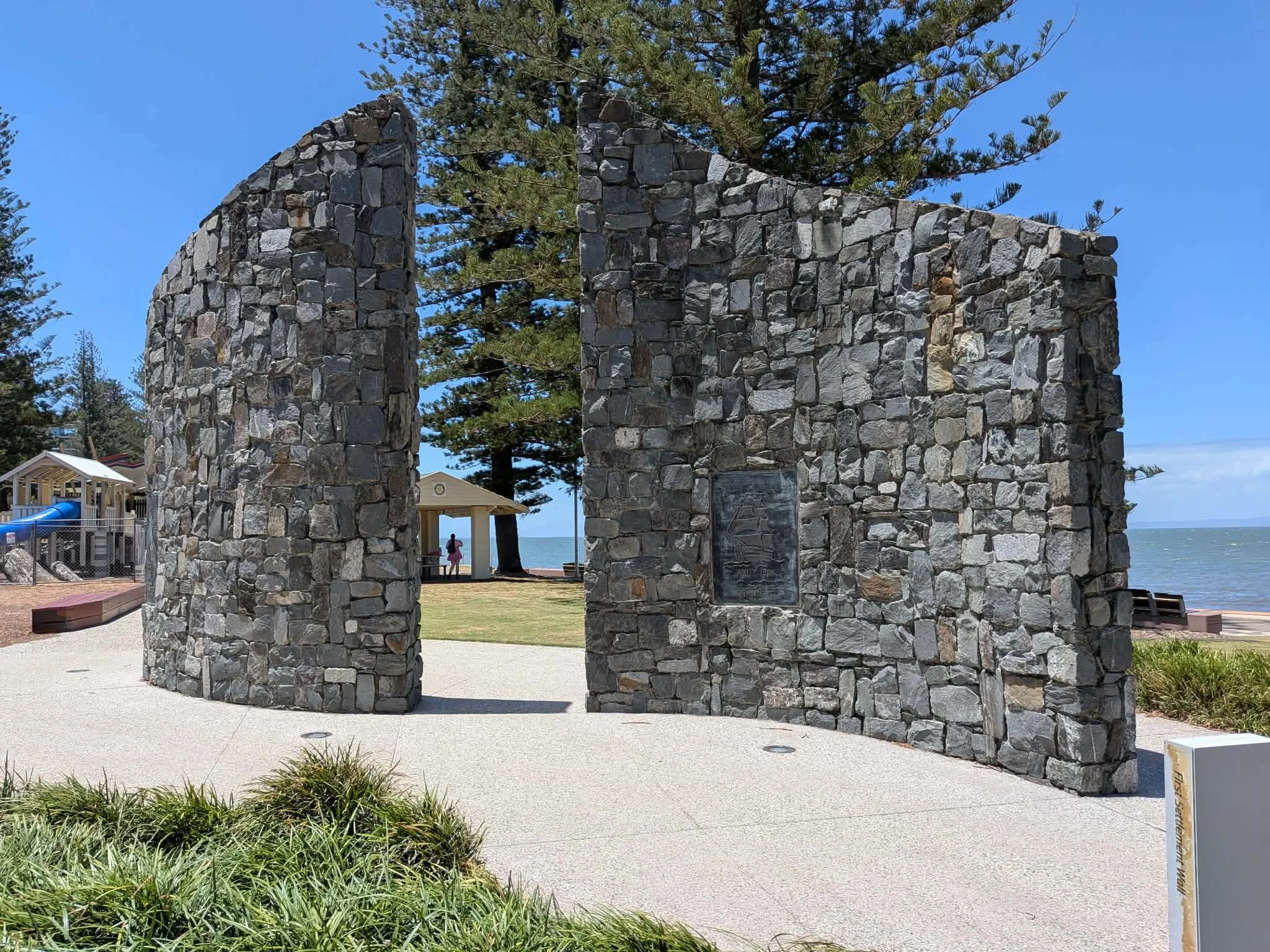 A large curved stone monument with a plaque stands near the beach, with trees, a playground, and a pavilion visible in the background, overlooking the sea. Redcliffe, Beach farewell, Funeral on the beach.