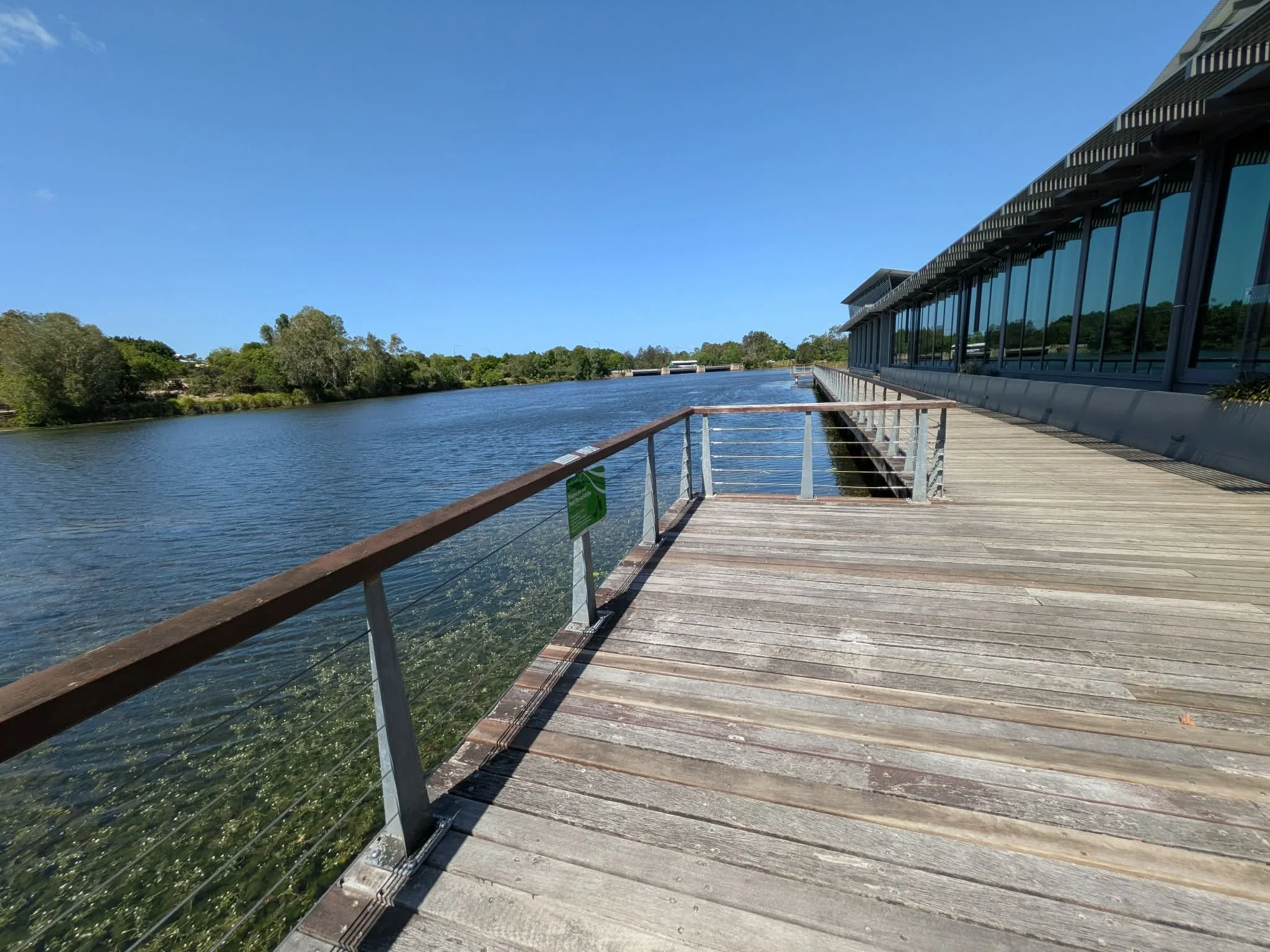 A wooden boardwalk along a river with a modern building on the right, blue sky, and green trees on the left. North Lakes.