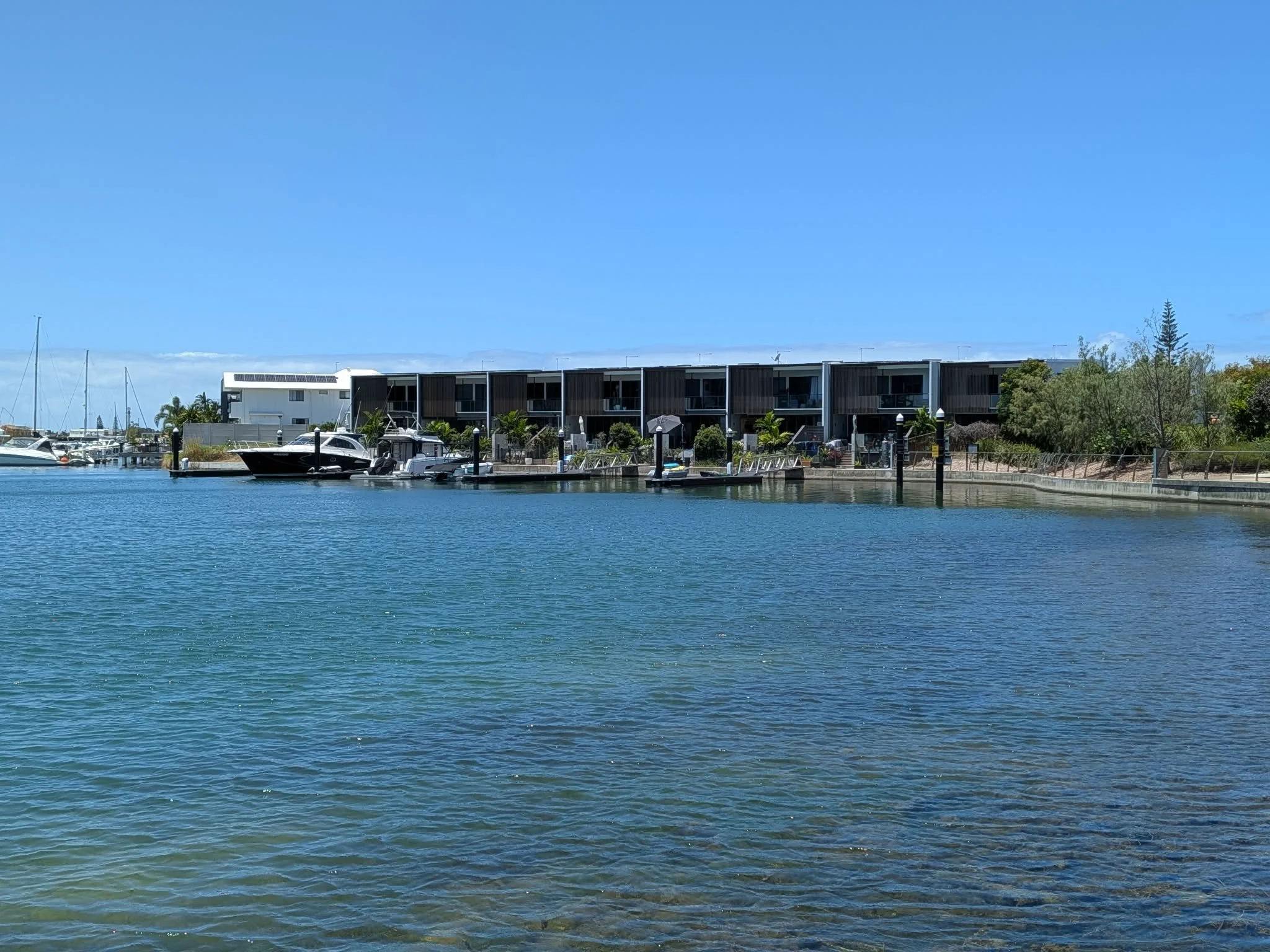 A marina with boats docked along the water, modern multi-story residential buildings in the background, greenery, and a clear blue sky. Beautiful place for a farewell service, Moreton Bay Memorial service.