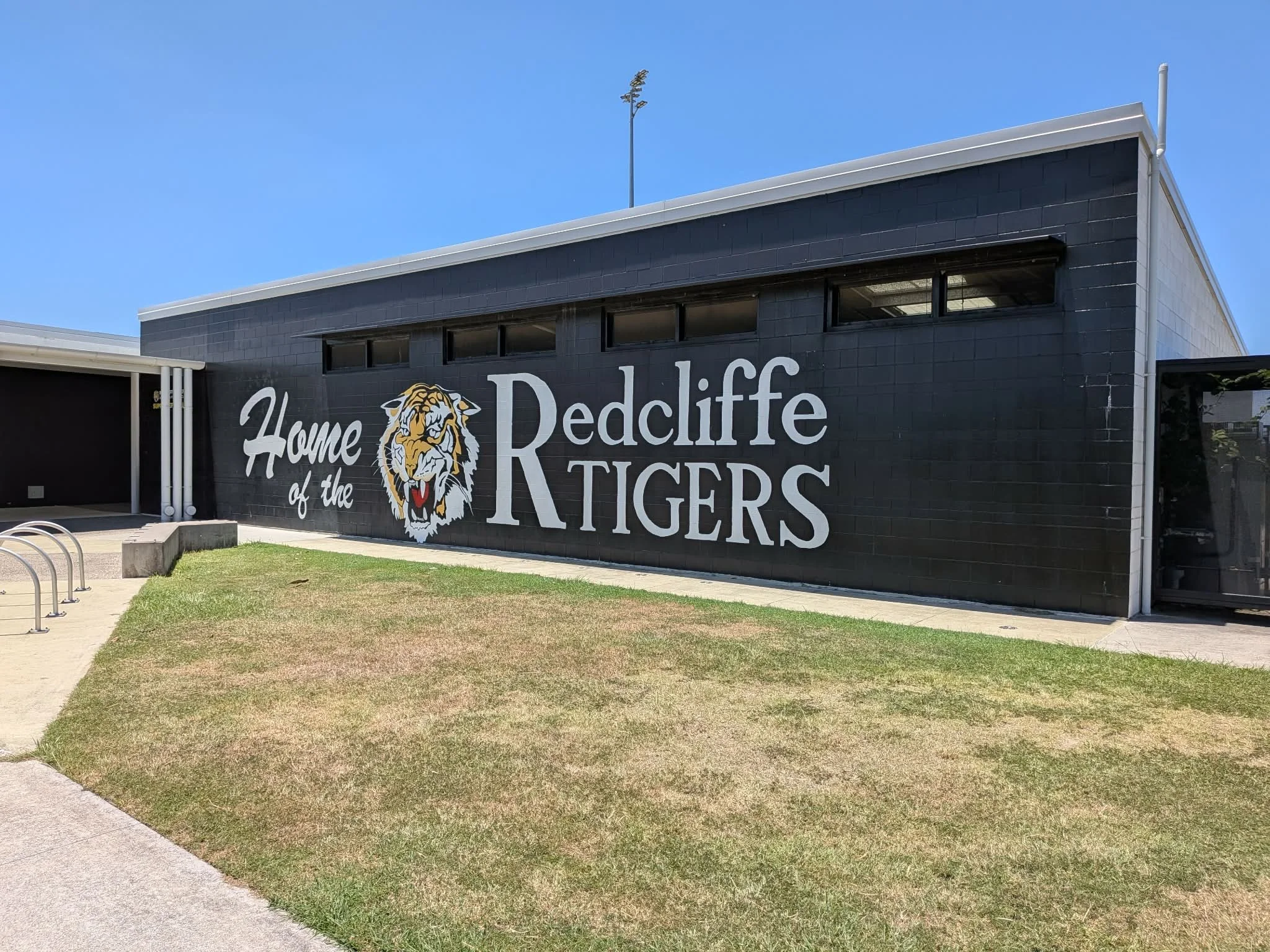 Black building with large sign reading "Home of the Redcliffe Tigers," featuring a tiger mascot, on a grassy area with sidewalk and bike racks in the foreground, under a clear blue sky. Saying farewell at their favourite sports club.