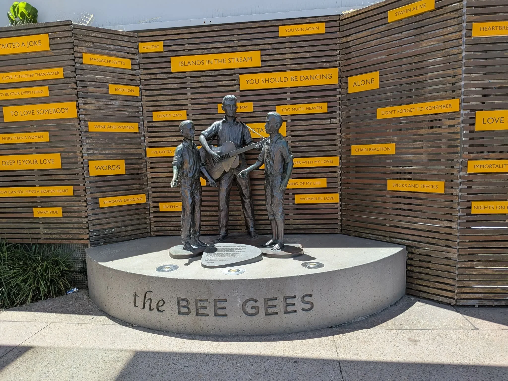 A statue of three band members from The Bee Gees playing instruments, set on a circular base with the text 'the BEE GEES', surrounded by a wooden wall with yellow plaques of song titles. Redcliffe.