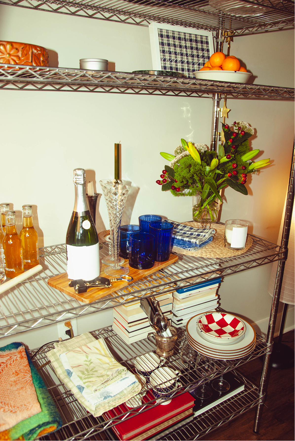 A metal kitchen shelf filled with various decorative and functional items, including a bouquet of flowers, stacks of plates, glasses, books, and a champagne bottle with glasses.