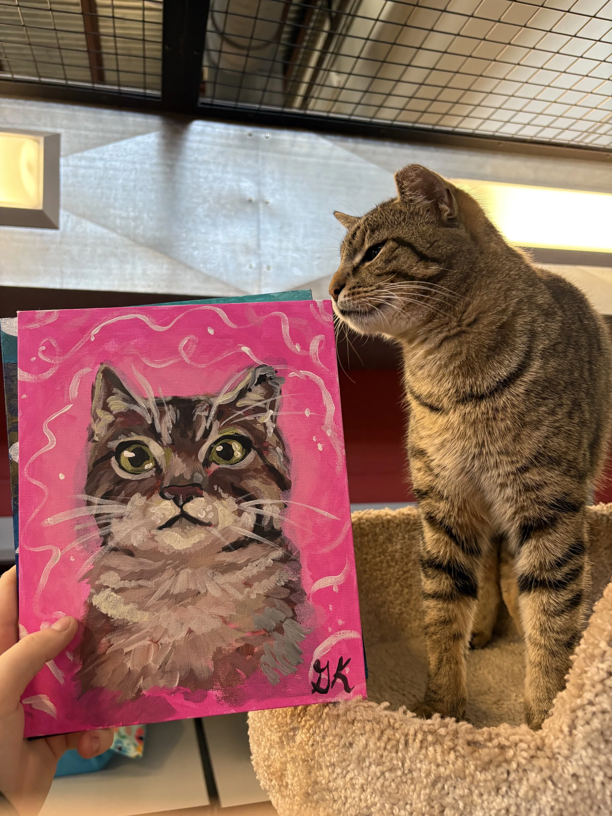 A person holds up a painting of a tabby cat with big green eyes and a pink background, next to a real tabby cat sitting in a beige plush bed.