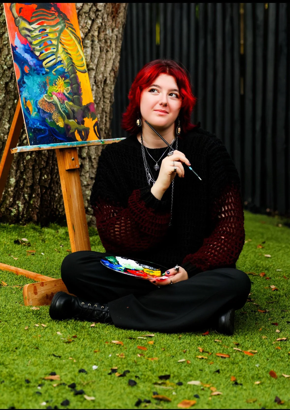 A woman with red hair sitting cross-legged on a grassy lawn, holding a paintbrush and palette, with a canvas of colorful abstract artwork and a tree trunk behind her.