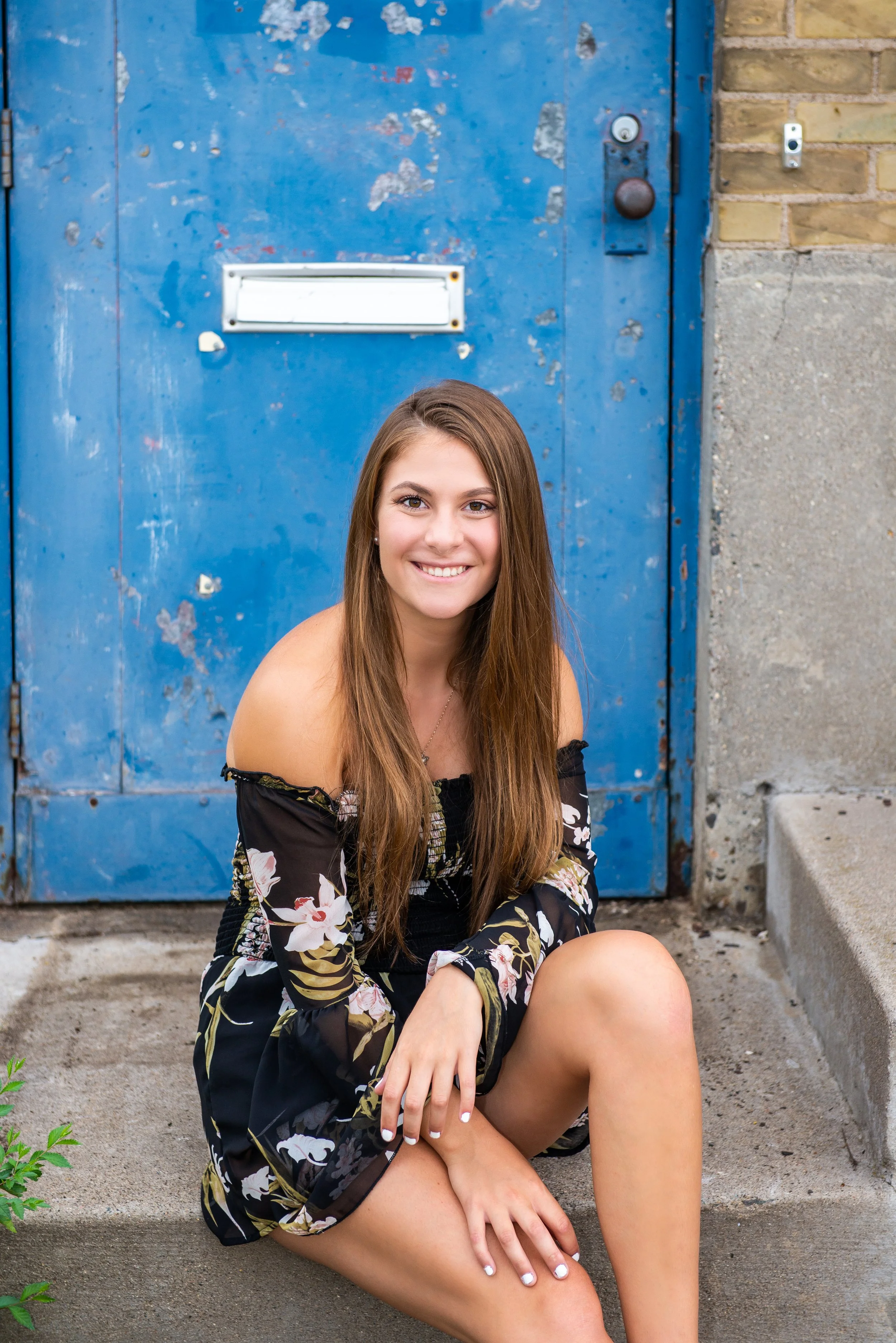 A young woman with long brown hair sitting on a concrete sidewalk in front of a weathered blue door, smiling at the camera.
