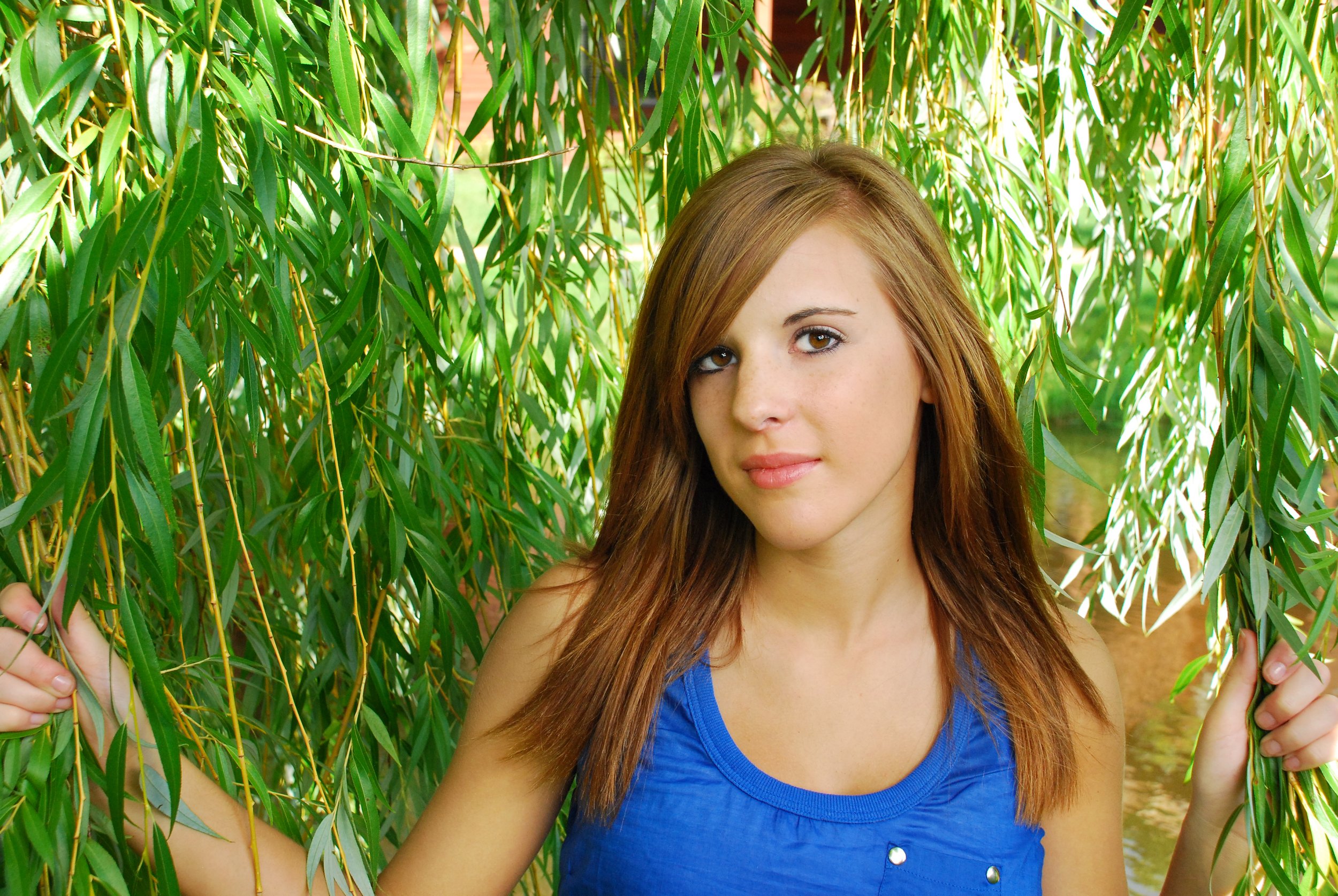A young woman with light skin and shoulder-length reddish-brown hair, wearing a blue sleeveless top, standing among green willow tree branches, holding onto the branches on each side.