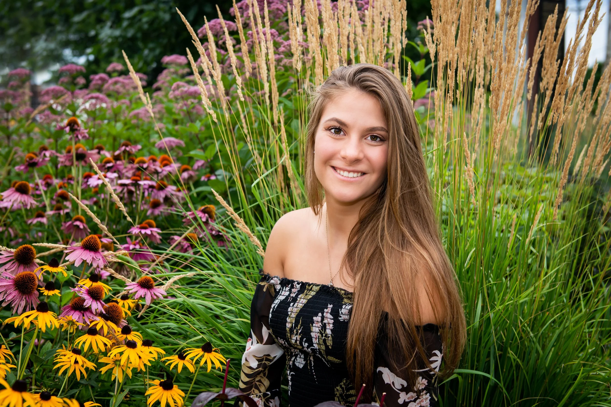 A young woman with long brown hair smiling in a garden with colorful flowers and tall grasses.
