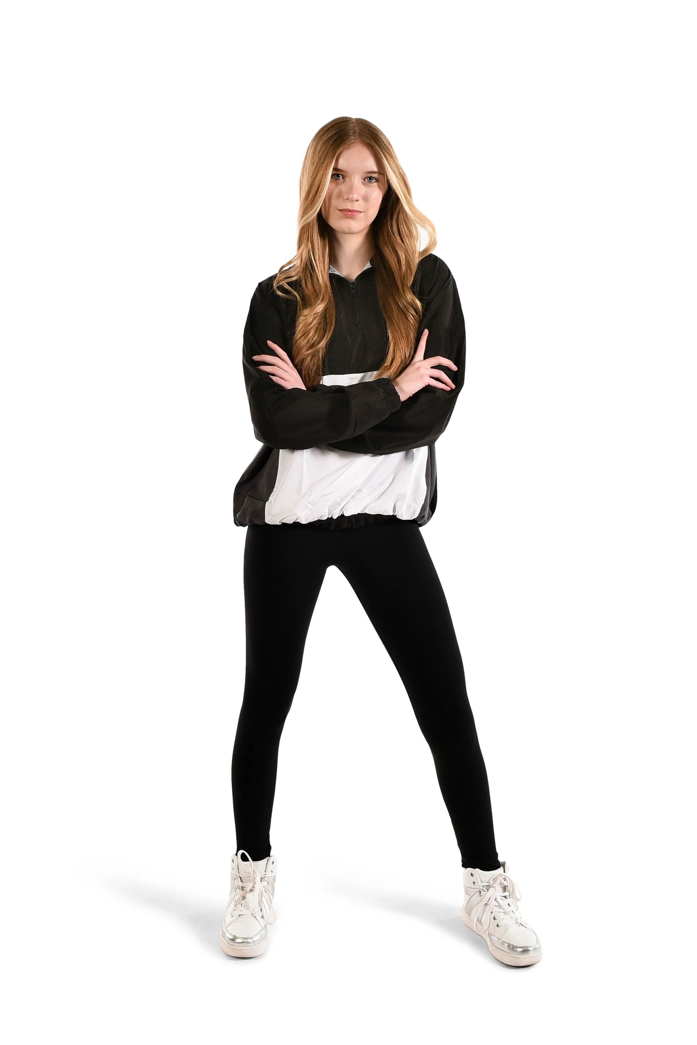 A young woman with long, wavy red hair, wearing a black and white windbreaker, black leggings, and white sneakers, standing with arms crossed against a white background.