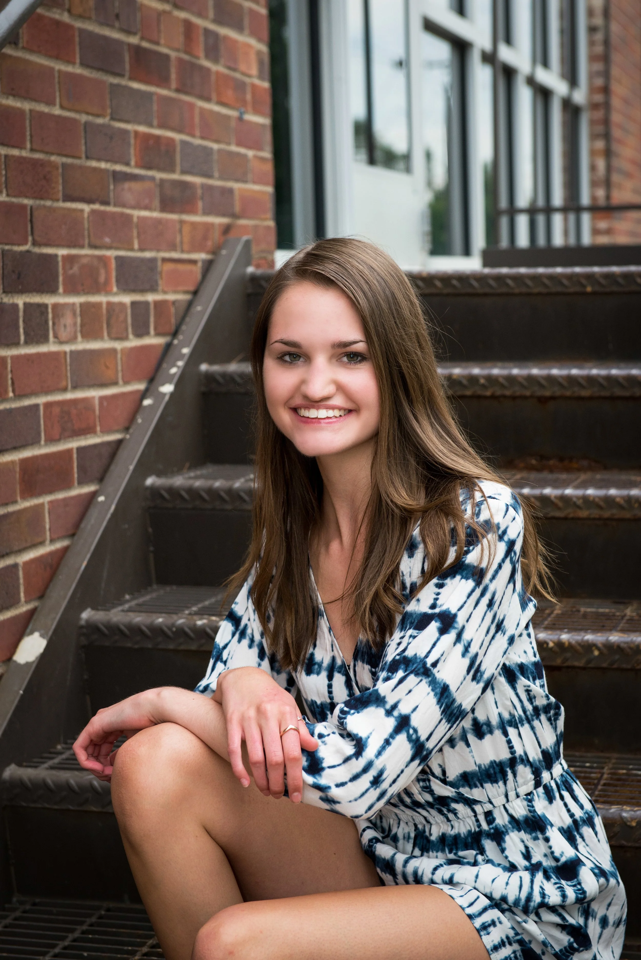 A young woman with long brown hair, wearing a blue and white tie-dye dress, sitting on outdoor metal stairs next to a brick building, smiling at the camera.