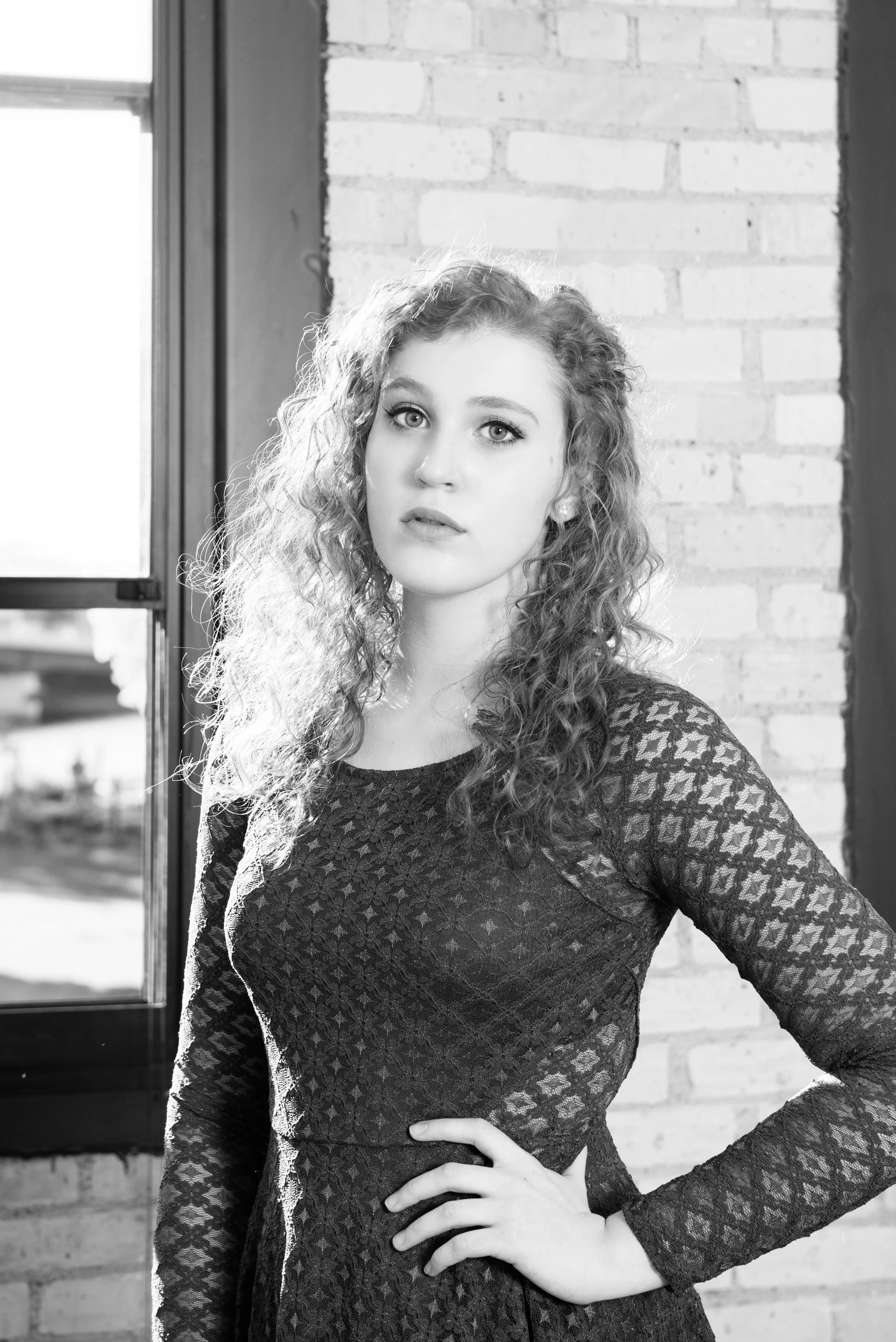Black and white photo of a young woman with curly hair standing indoors near a window and brick wall, wearing a lace dress with one hand on her hip.