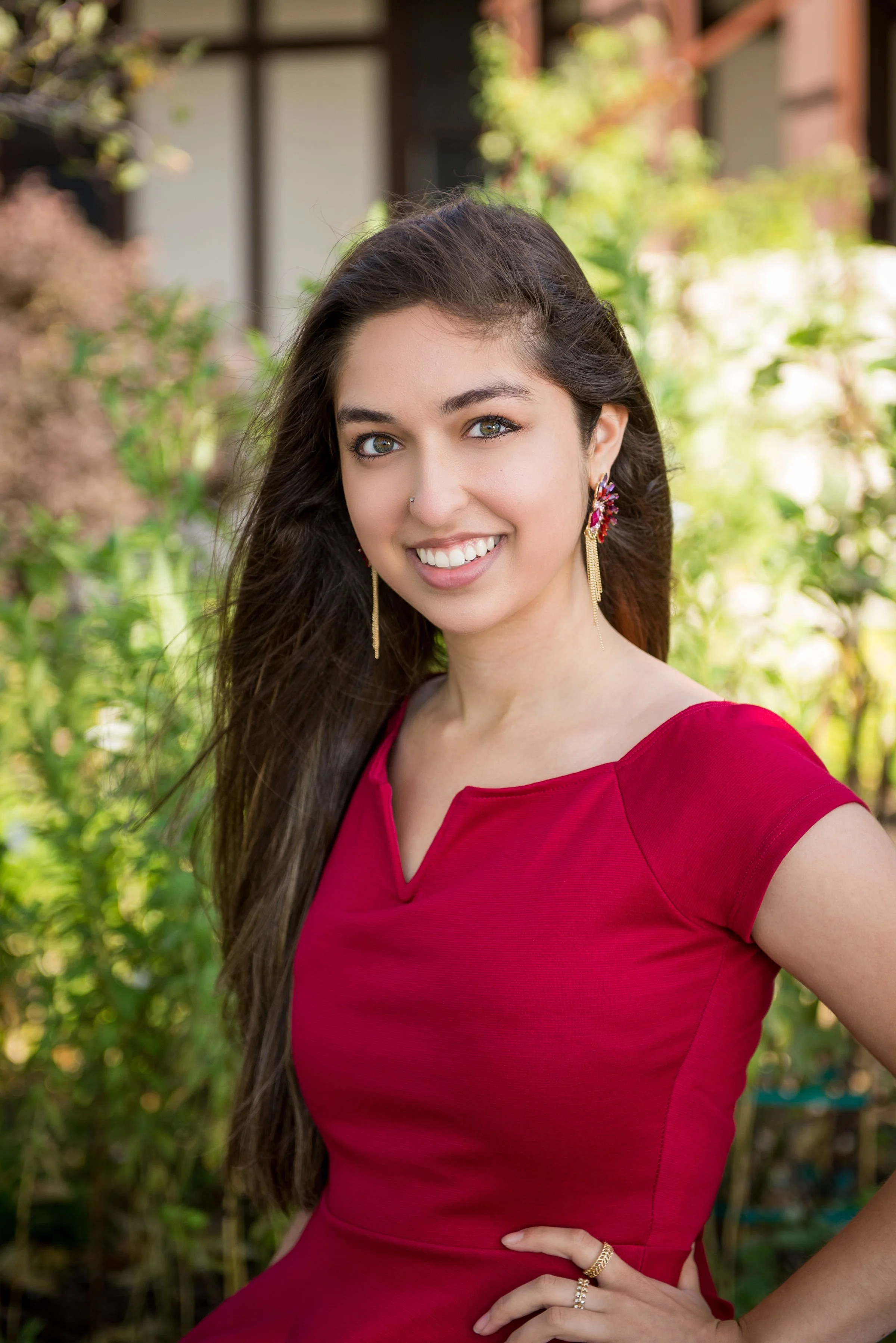 A young woman with long dark hair, wearing a red dress, smiling outdoors with green plants in the background.