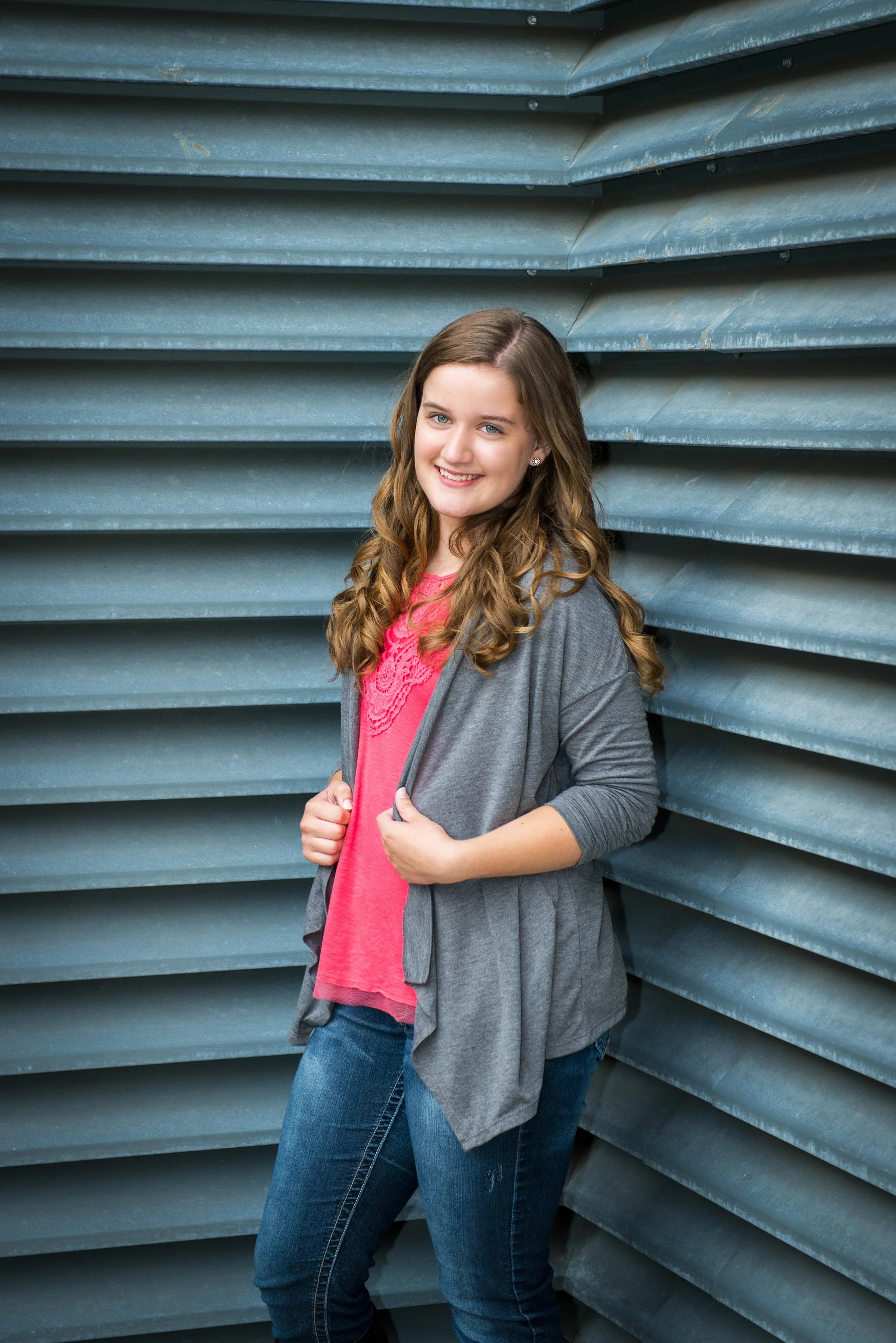 A young woman with long, curly brown hair smiling while standing against a modern, corrugated metal wall. She is wearing a pink top, a gray cardigan, and blue jeans.