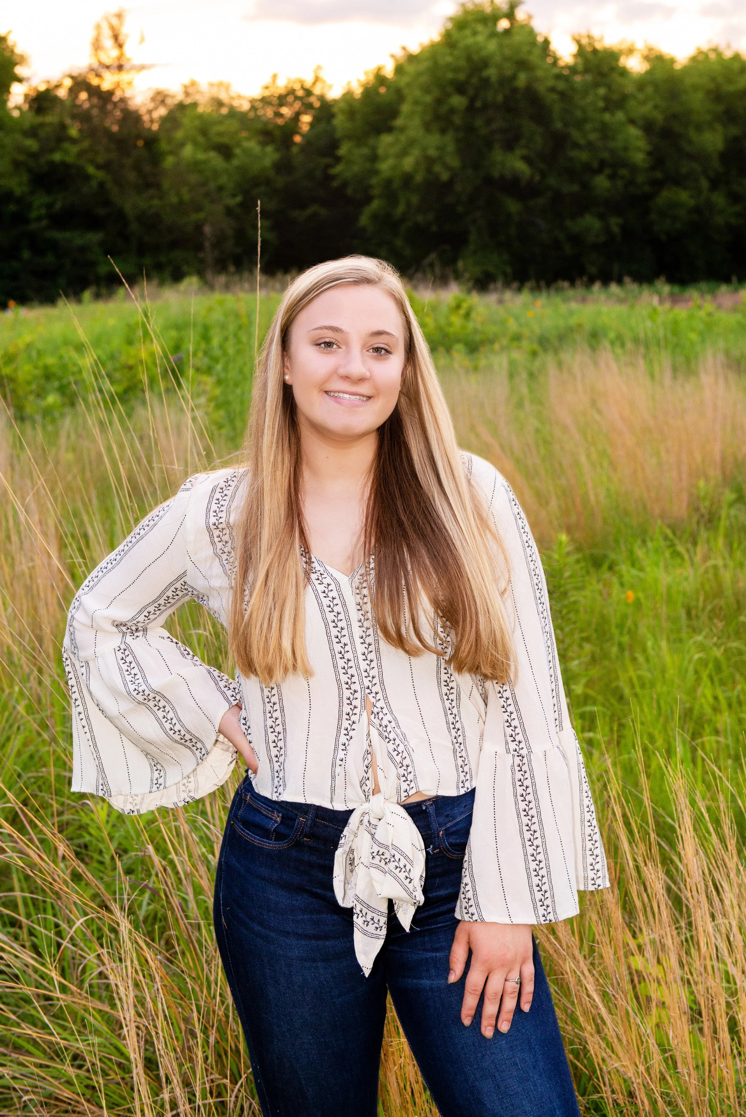 A young woman stands in a grassy field at sunset, smiling with one hand on her hip and wearing a white blouse with black pattern and dark jeans.