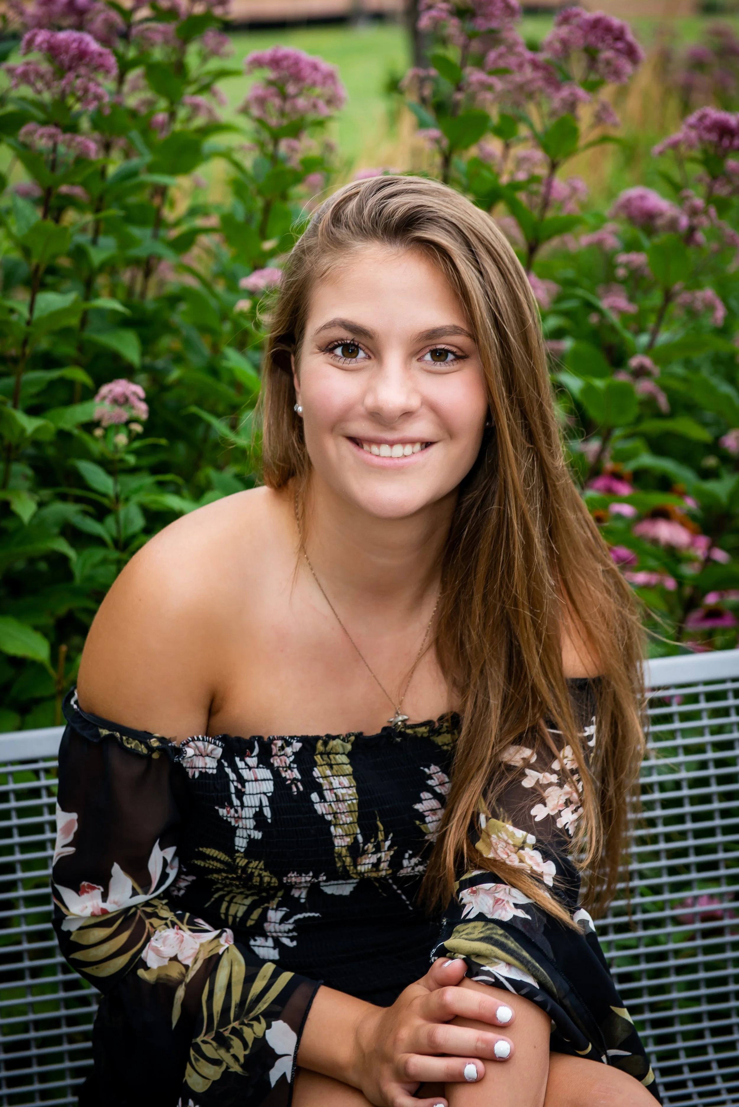 A young woman with long brown hair smiling, sitting on a metal bench outdoors in front of pink flowering plants.